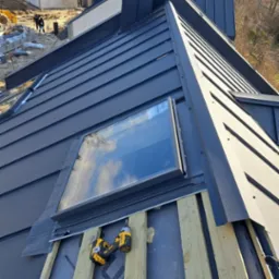 Montaż okna dachowego na kryciu z blachy na rąbek stojący. Na połaci dachu leżą dwa wkrętaki akumulatorowe. W tle budynki w trakcie budowy.