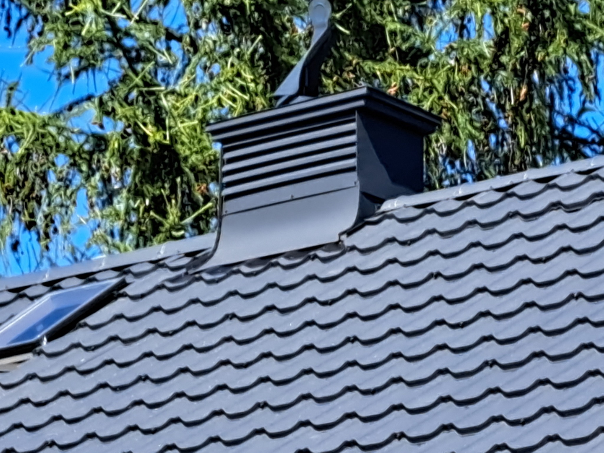 Szary dach z dachówki ceramicznej z czarnym kominem i lukarną, na tle drzewa w Raciążu. Widok z bliska, oświetlenie dzienne.