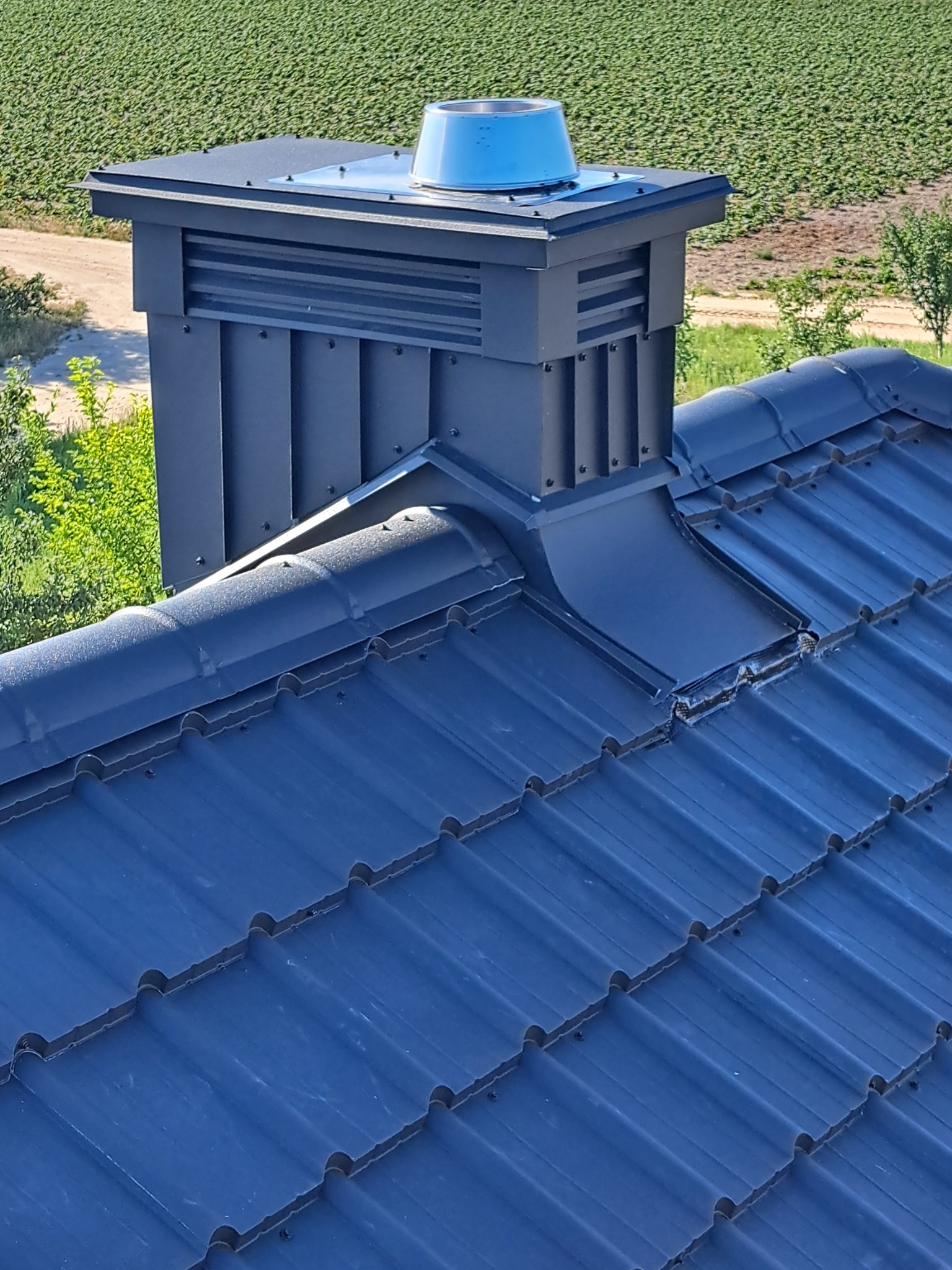 Ciemny, metalowy komin wentylacyjny na dachu z ciemnej dachówki ceramicznej. W tle pole i drzewa. Widok z góry.