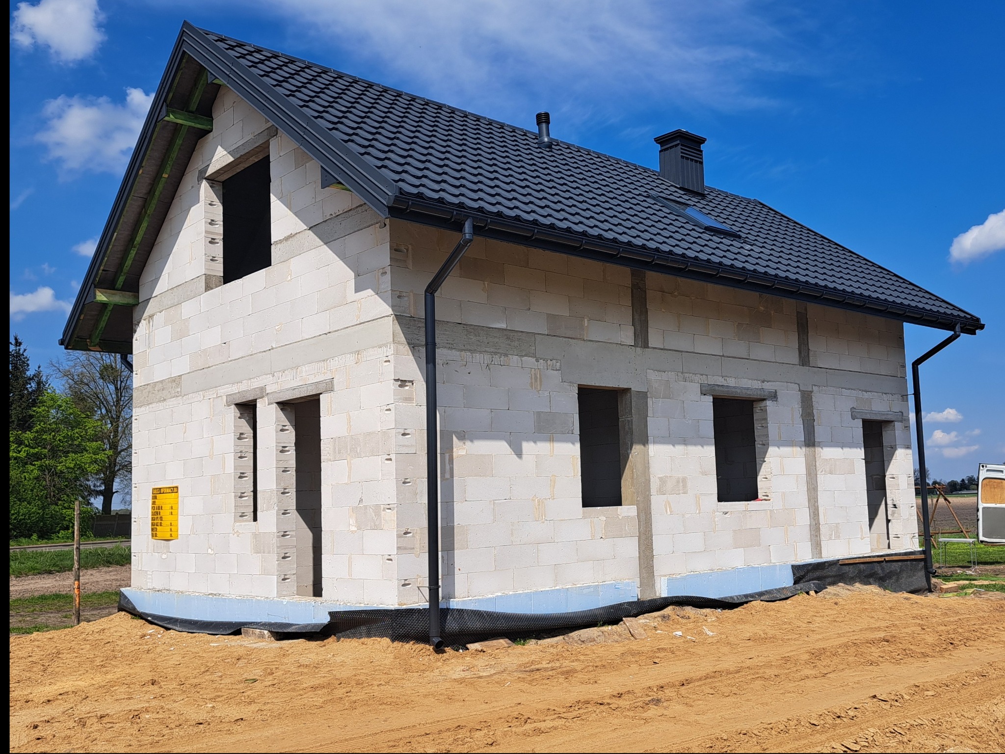 Surowy dom z czarnym dachem z dachówki, widoczne rynny, okna bez szyb, na ścianie żółta tablica informacyjna, teren wokół domu piaszczysty.