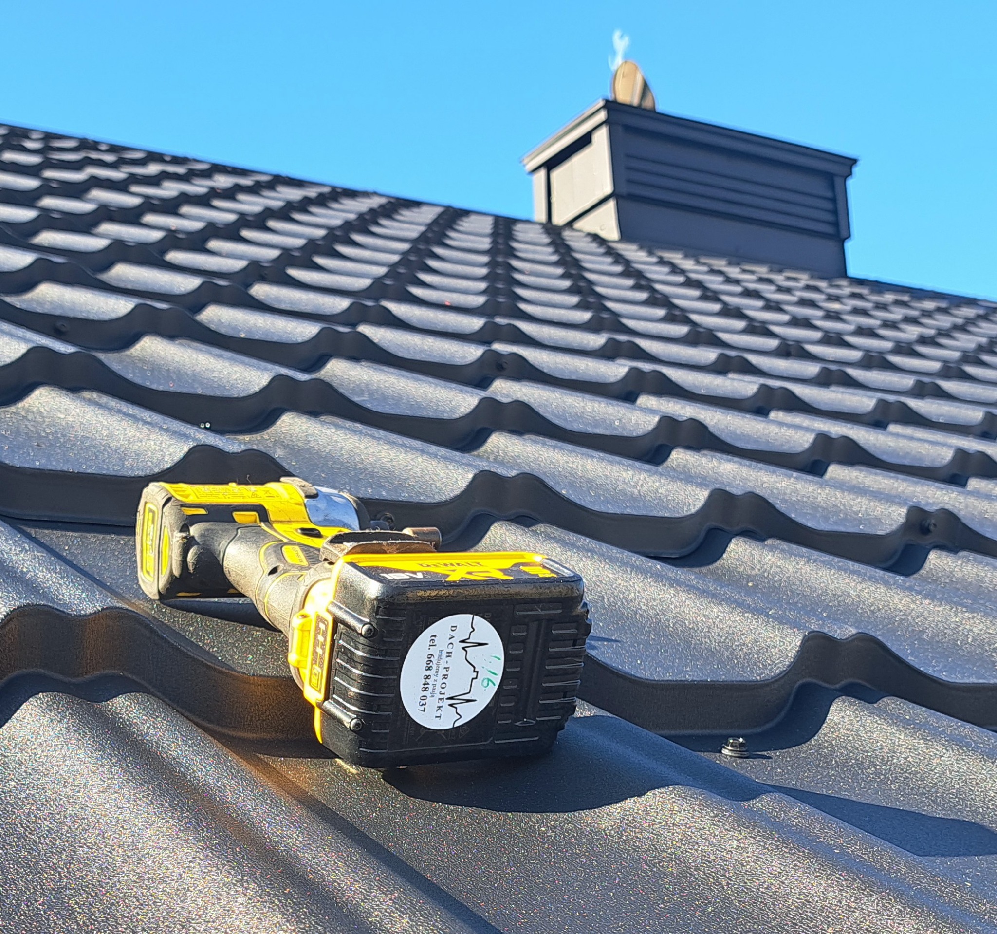 Czarny dach z blachodachówki modułowej, na nim wkrętarka akumulatorowa z naklejką firmy dekarskiej. W tle komin i błękitne niebo.