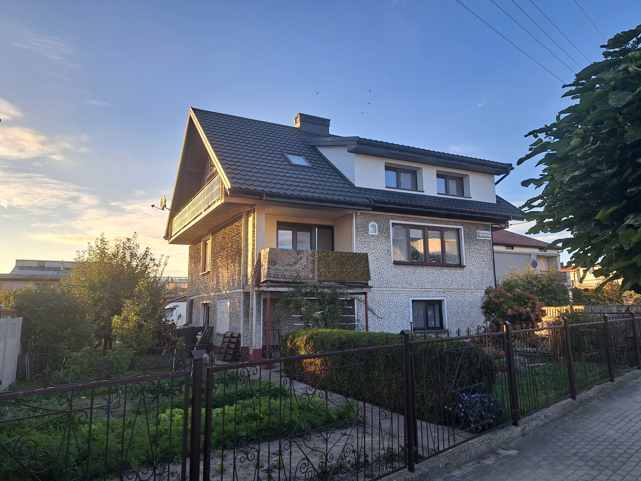 Dwupiętrowy dom z ciemnym dachem i balkonami, widziany zza metalowego ogrodzenia ogrodu, w ciepłym świetle słonecznym. Widoczne okna dachowe i komin.