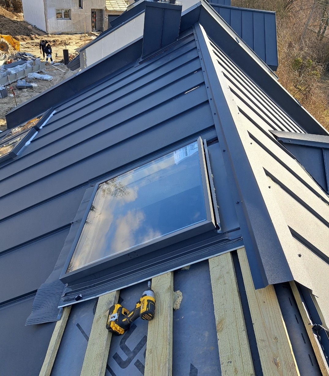 Montaż okna dachowego na kryciu z blachy na rąbek stojący. Na połaci dachu leżą dwa wkrętaki akumulatorowe. W tle budynki w trakcie budowy.