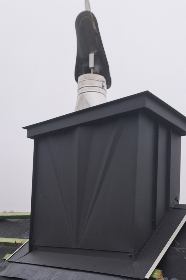 Nowoczesny, czarny komin z metalowym wykończeniem na dachu. Widoczne detale obróbki blacharskiej i elementy wentylacyjne. Ujęcie z dołu, pochmurne niebo w tle.