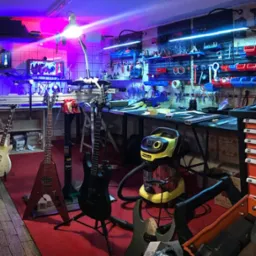 Warsztat naprawy gitar z narzędziami i instrumentami. Uwagę przykuwa żółty odkurzacz Karcher oraz logotyp firmy 'Hammer-On Guitar Service'.