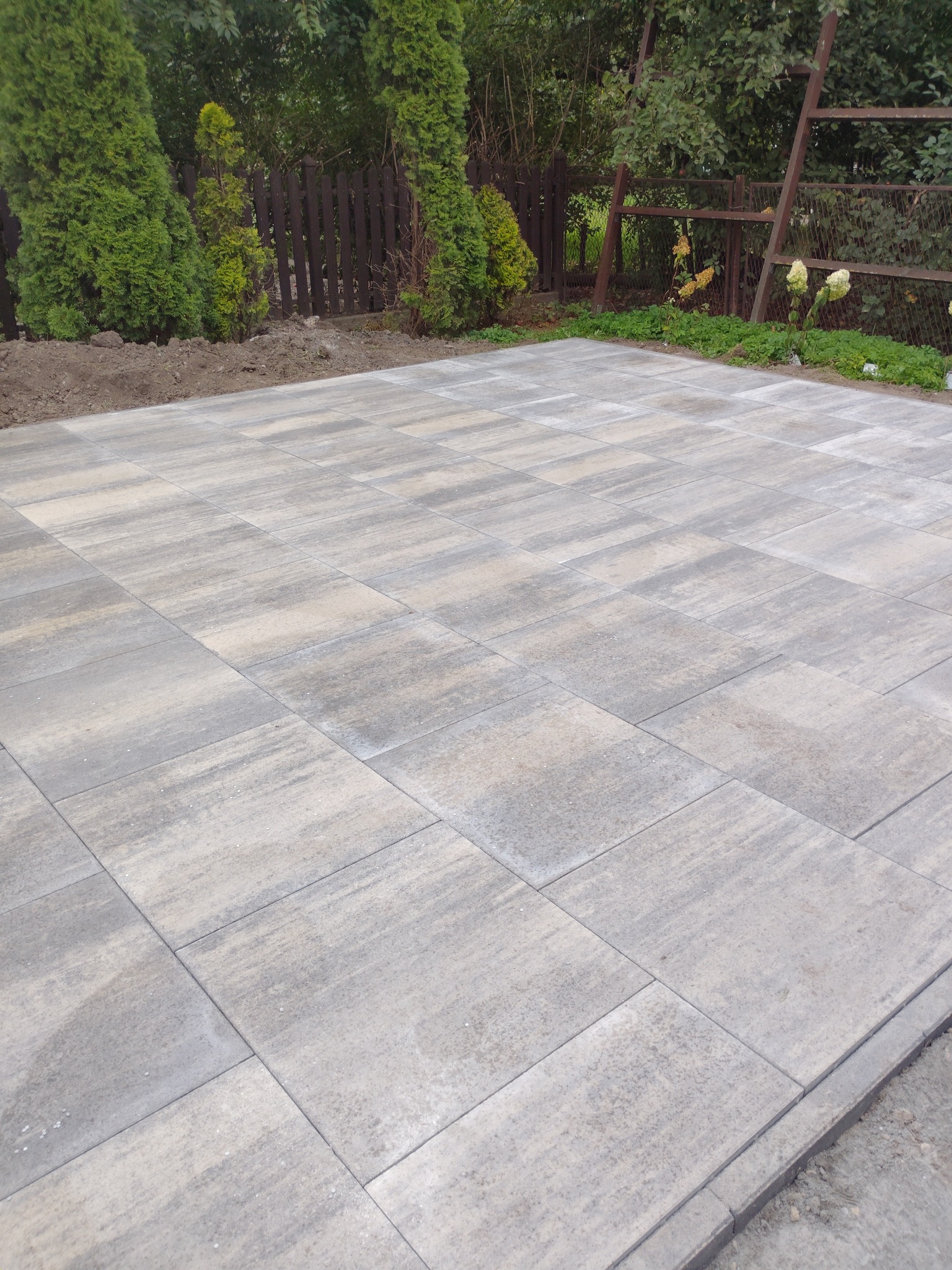 Ułożona, szara kostka brukowa na posesji, widok z góry. W tle roślinność i drewniany płot. Równa powierzchnia, schludne wykończenie.