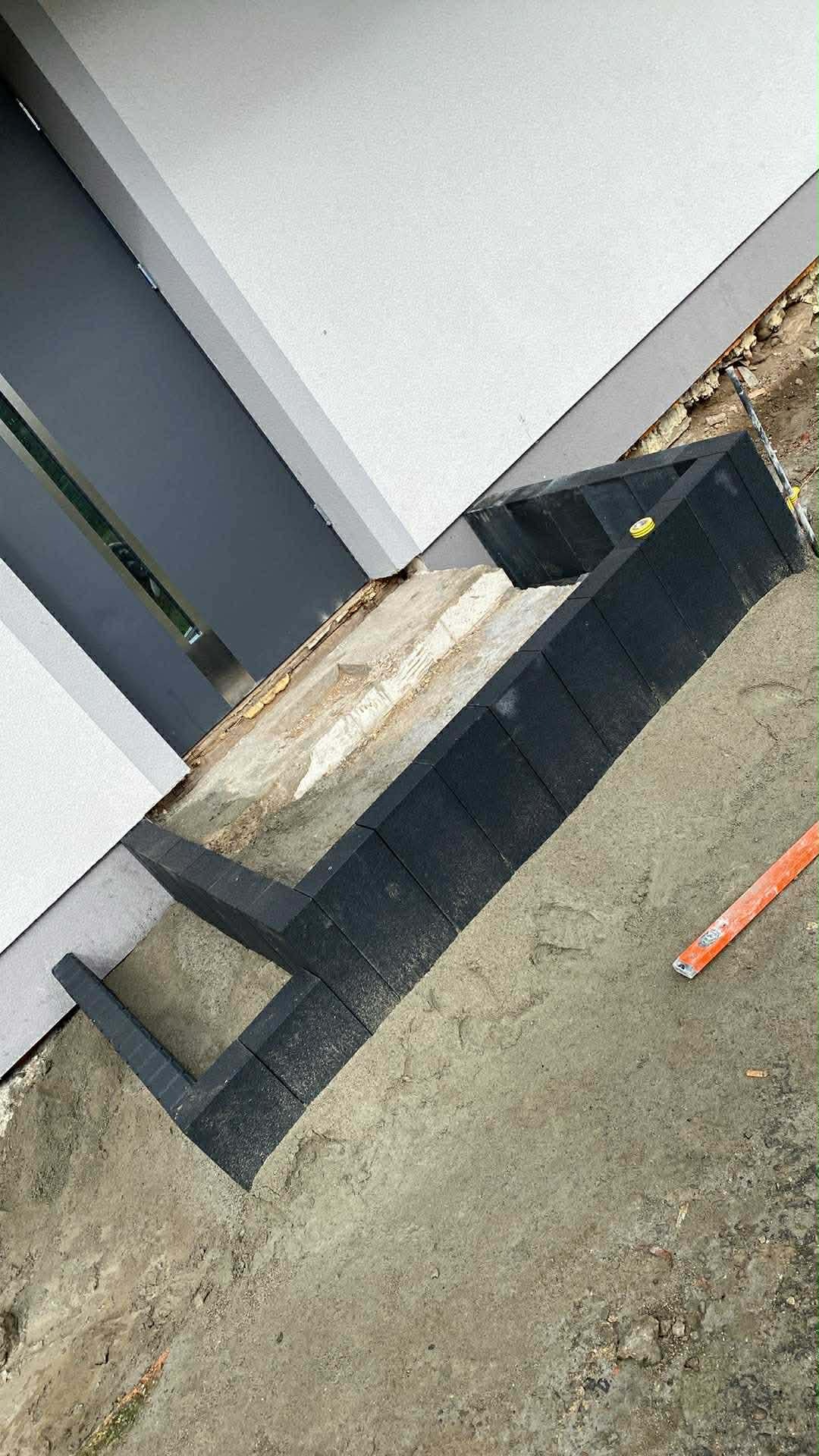 Układanie obrzeży z czarnej kostki brukowej przy budynku, widoczna poziomica. Prace brukarskie w toku, przygotowanie podłoża. Kątowe ułożenie kostki.