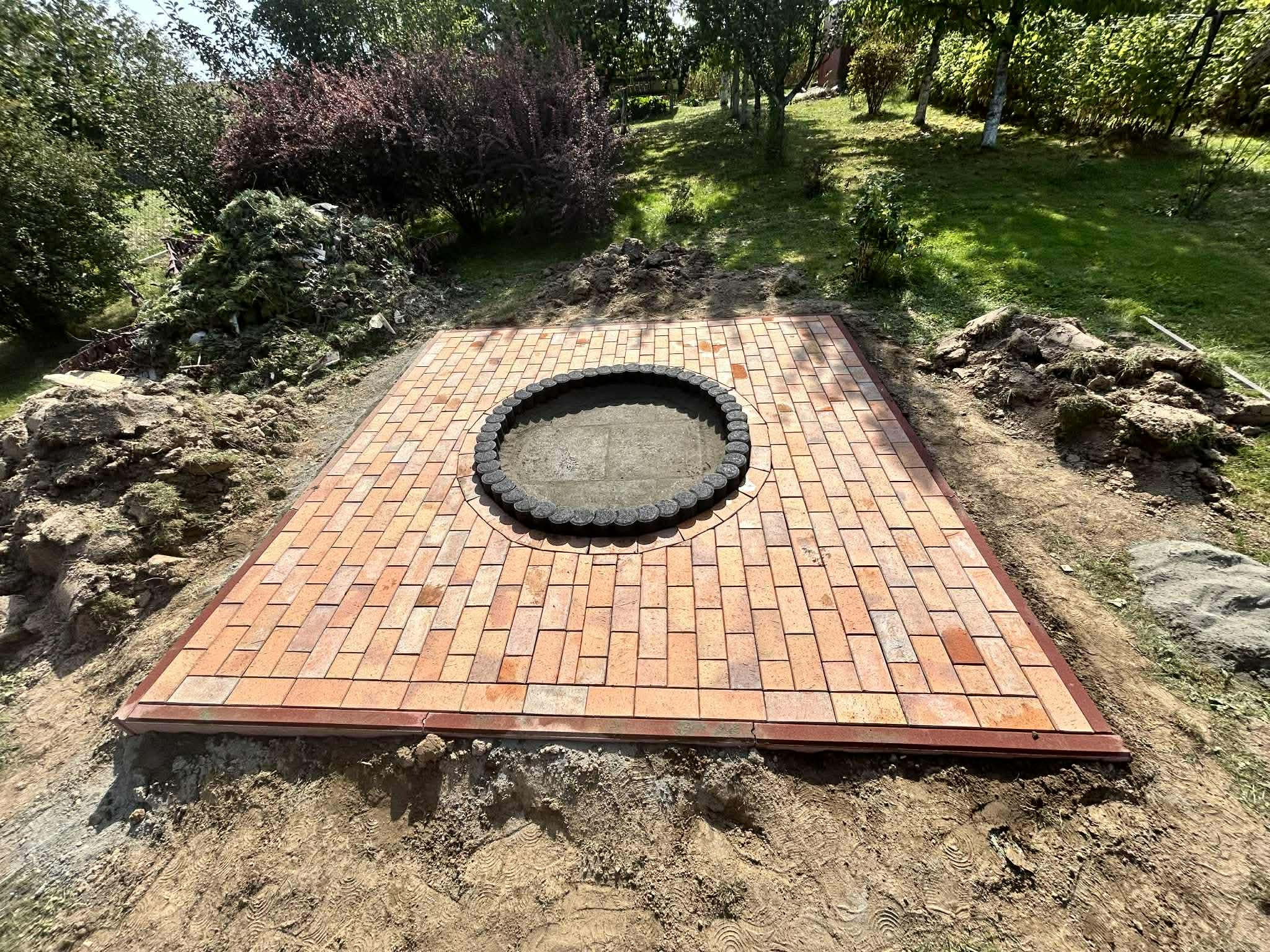 Kwadratowa powierzchnia z ceglanej kostki z okrągłym paleniskiem pośrodku, otoczona ziemią i zielenią. Widok z góry, ciepłe światło słoneczne.