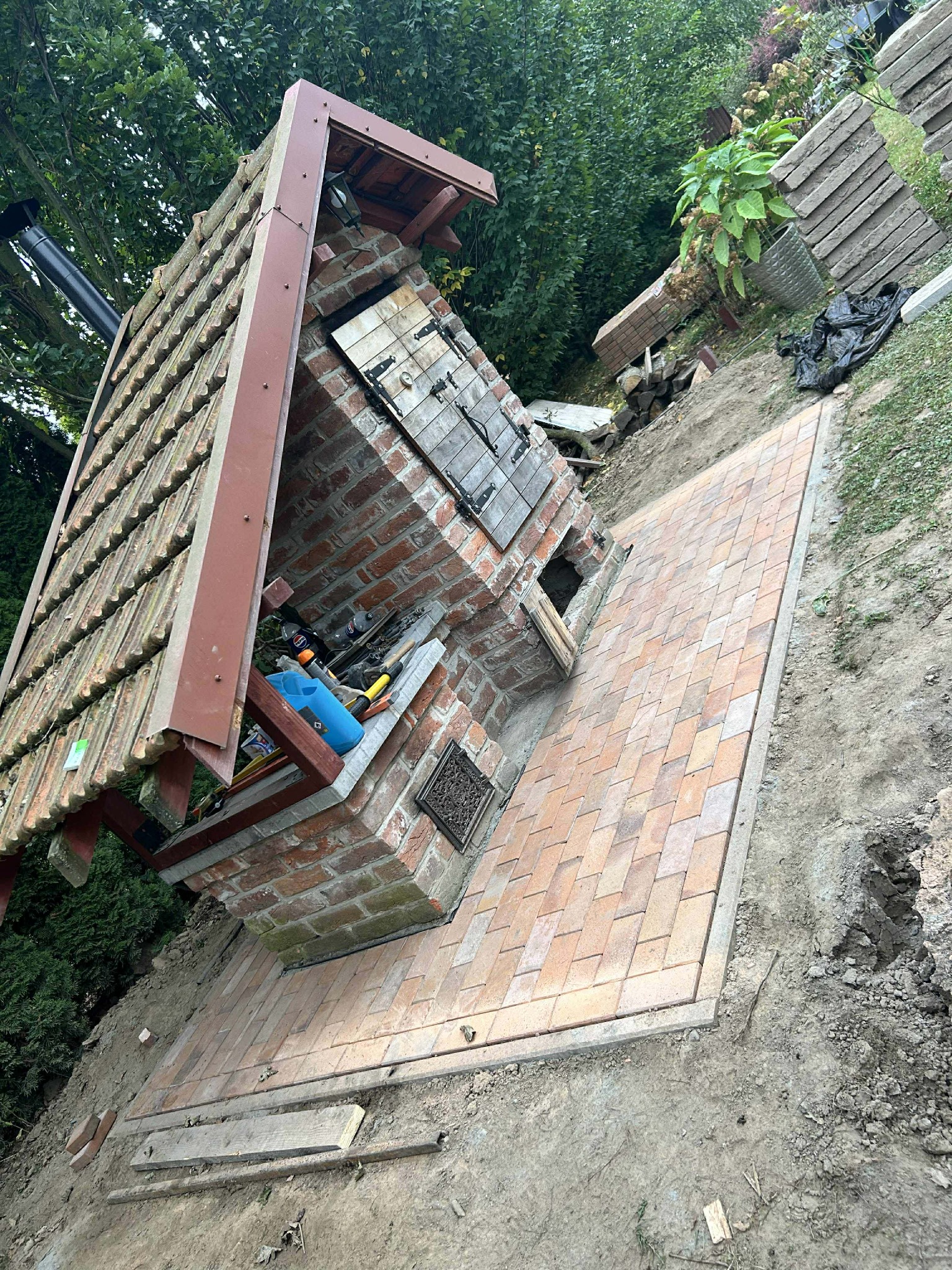Ukończona ścieżka z kostki prowadząca do ceglanego grilla z zadaszeniem, otoczona ziemią i zielenią. Widoczne narzędzia i materiały budowlane.