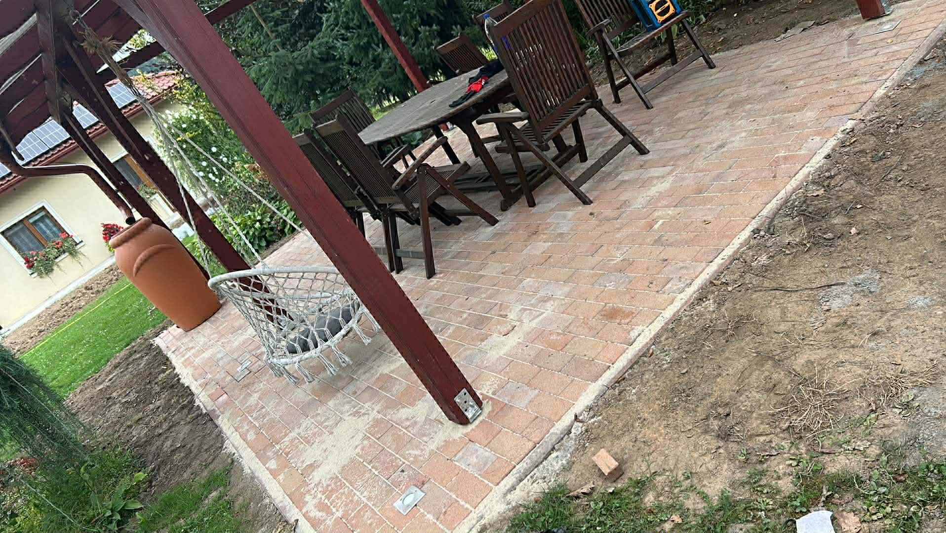 Ceglana kostka na tarasie z drewnianą pergolą, huśtawką i meblami ogrodowymi. Ułożona w jodełkę, z widocznymi śladami piasku. Otoczenie: trawnik i drzewa.