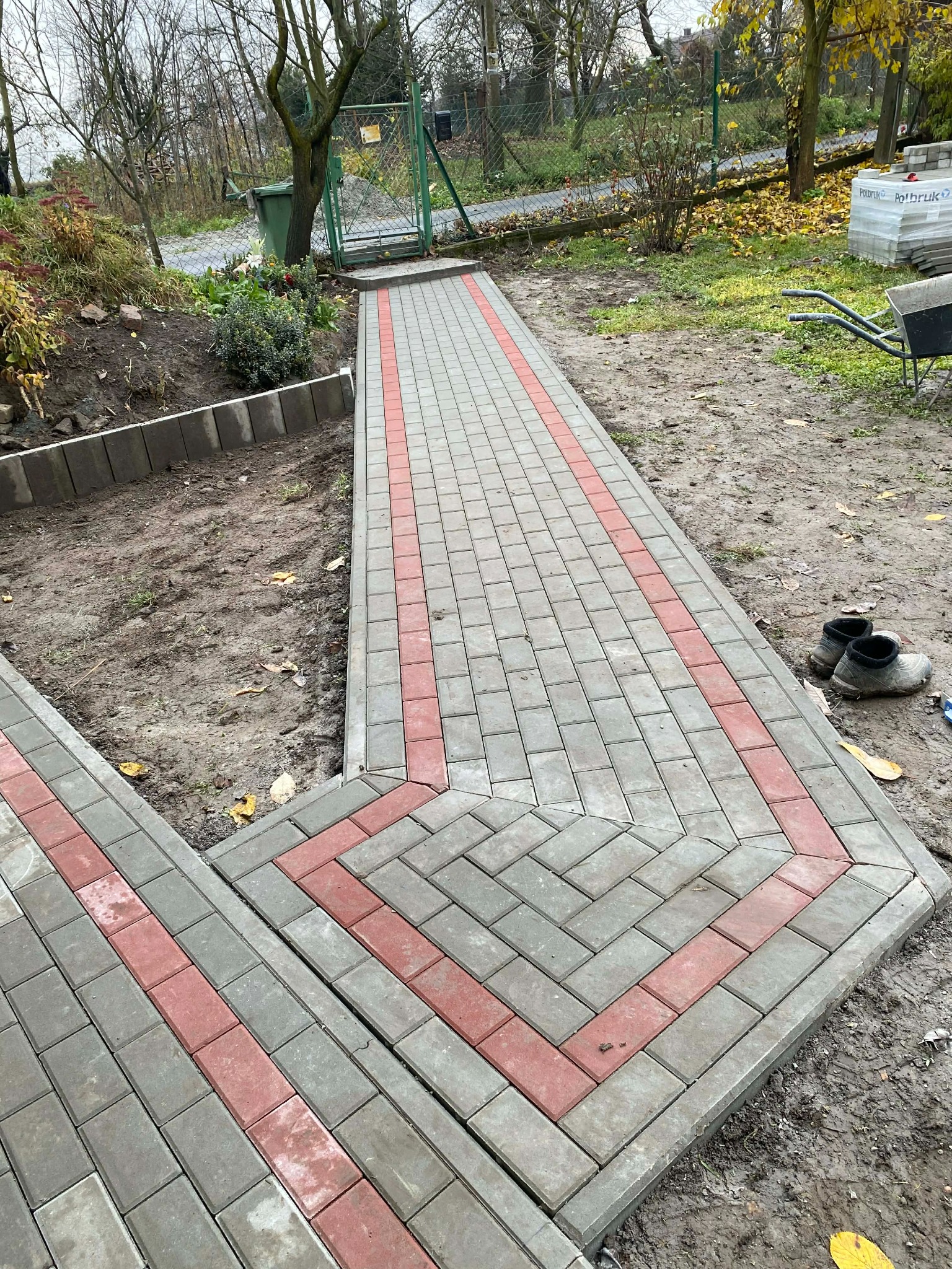 Ścieżka z kostki brukowej w odcieniach szarości i czerwieni, ułożona w geometryczny wzór. W tle widoczne drzewa i elementy ogrodu, buty robocze na kostce.