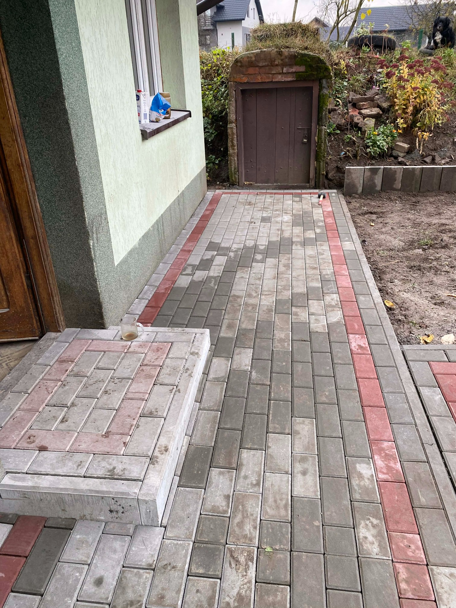 Ułożona kostka brukowa w odcieniach szarości i czerwieni przed budynkiem z zieloną elewacją i piwniczką ziemną. Widoczny fragment schodów z kostki.