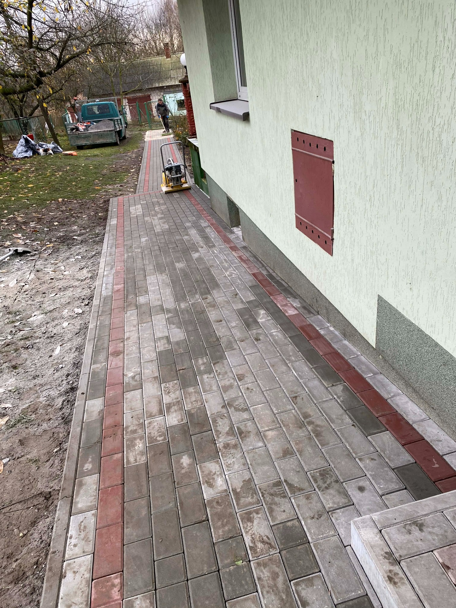 Ścieżka z kostki brukowej w odcieniach szarości i czerwieni, prowadząca wzdłuż ściany budynku. W tle widoczny pracownik z maszyną do ubijania kostki i samochód dostawczy.