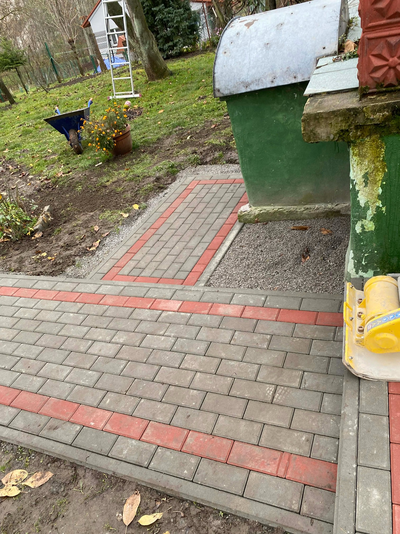 Ułożona ścieżka z kostki brukowej w odcieniach szarości i czerwieni, prowadząca do zielonego budynku. Żółta zagęszczarka w rogu kadru. Widok z góry.