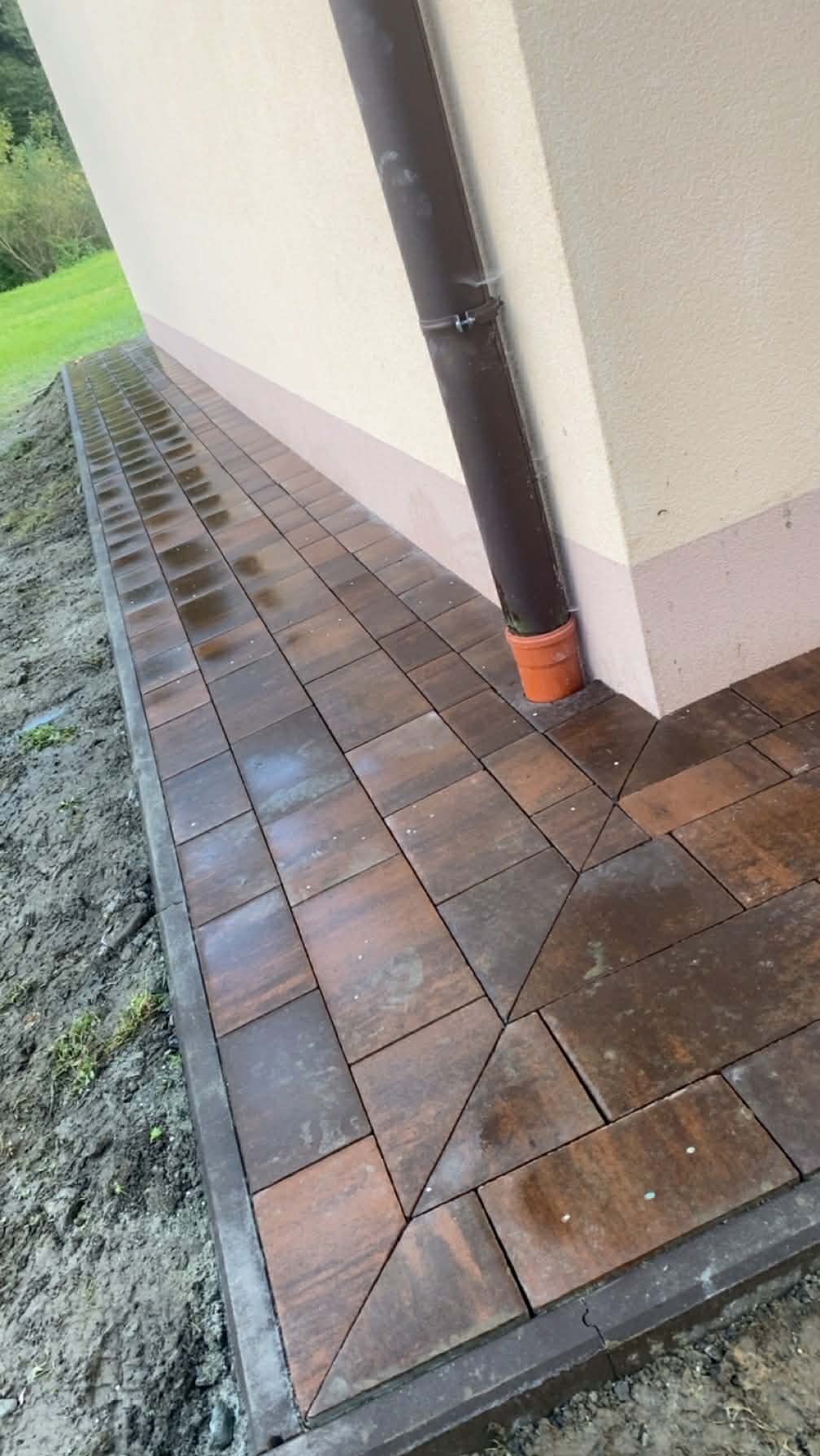 Ułożona, brązowa kostka brukowa przy ścianie budynku z rynną. Widoczne geometryczne wzory z kostki i obrzeża. Częściowo mokra nawierzchnia.