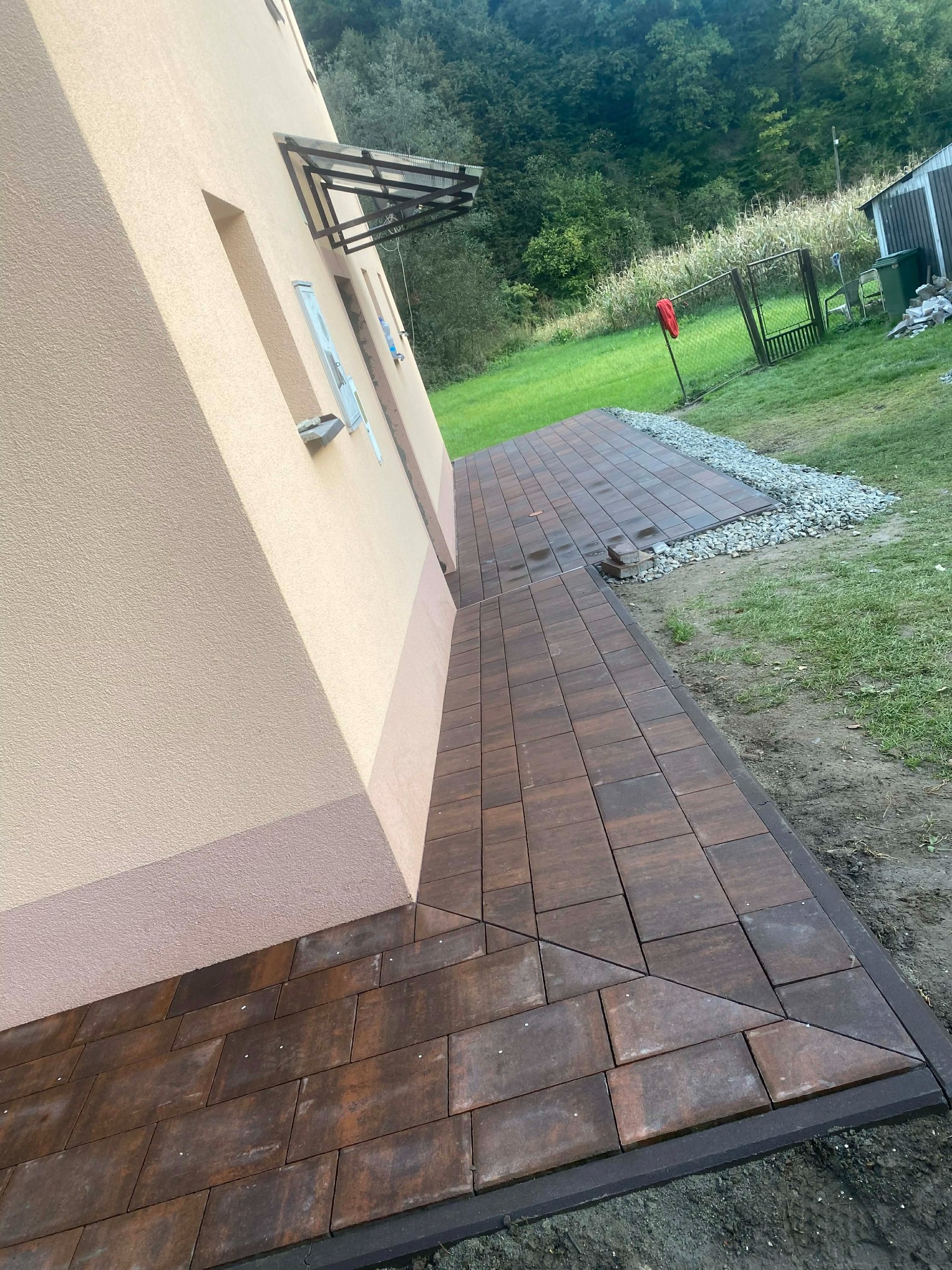 Brązowa kostka brukowa ułożona przy ścianie domu, tworząca ścieżkę. Widoczne elementy trawnika i ogrodu w tle. Kąt ujęcia podkreśla geometrię wzoru kostki.