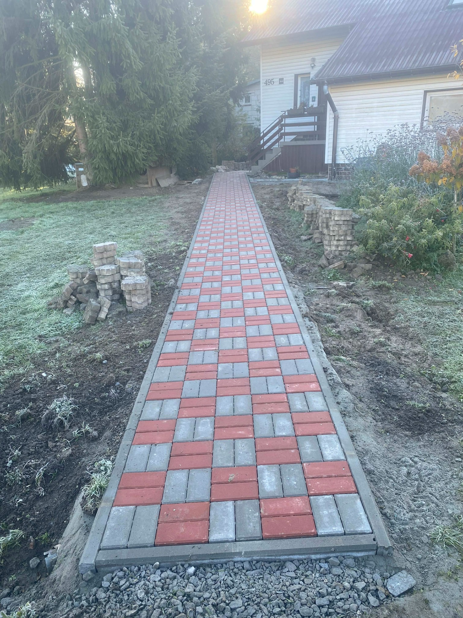 Ścieżka z kostki brukowej w szachownicę (czerwono-szara) prowadząca do domu. Ułożona kostka otoczona zielenią i stertami nieużytej kostki. Słoneczny dzień.