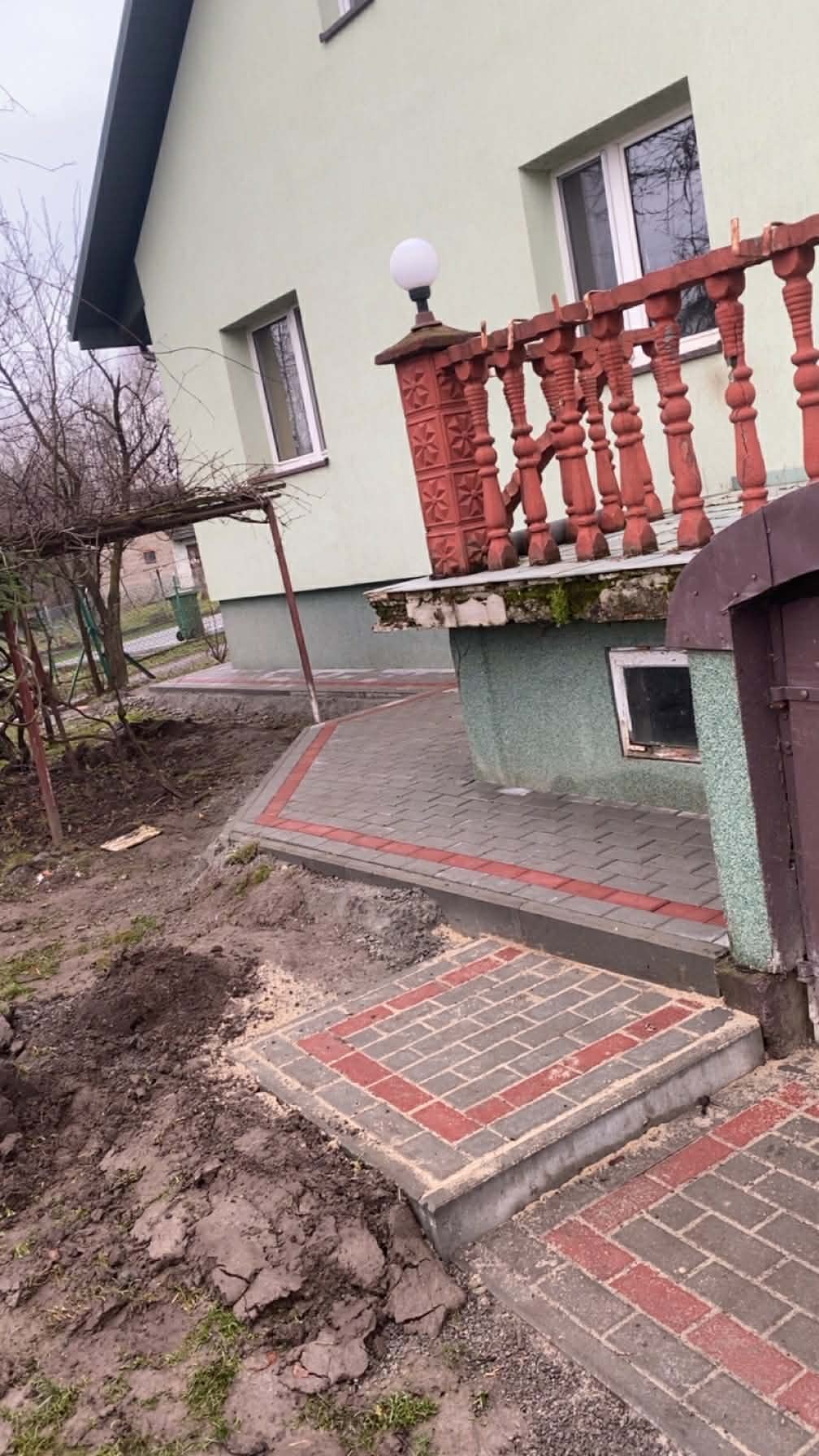 Ułożona kostka brukowa w odcieniach szarości i czerwieni tworzy ścieżkę i schody przed domem z zieloną elewacją i czerwonymi balustradami. Częściowo wykopana ziemia obok.