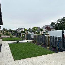 Mateusz Łucjan - Nowoczesny ogród z równo przystrzyżonym trawnikiem, świeżo posadzone drzewka podparte palikami, rabaty kwiatowe i szara palisada. Na podjeździe stoi biały samochód dostawczy.
