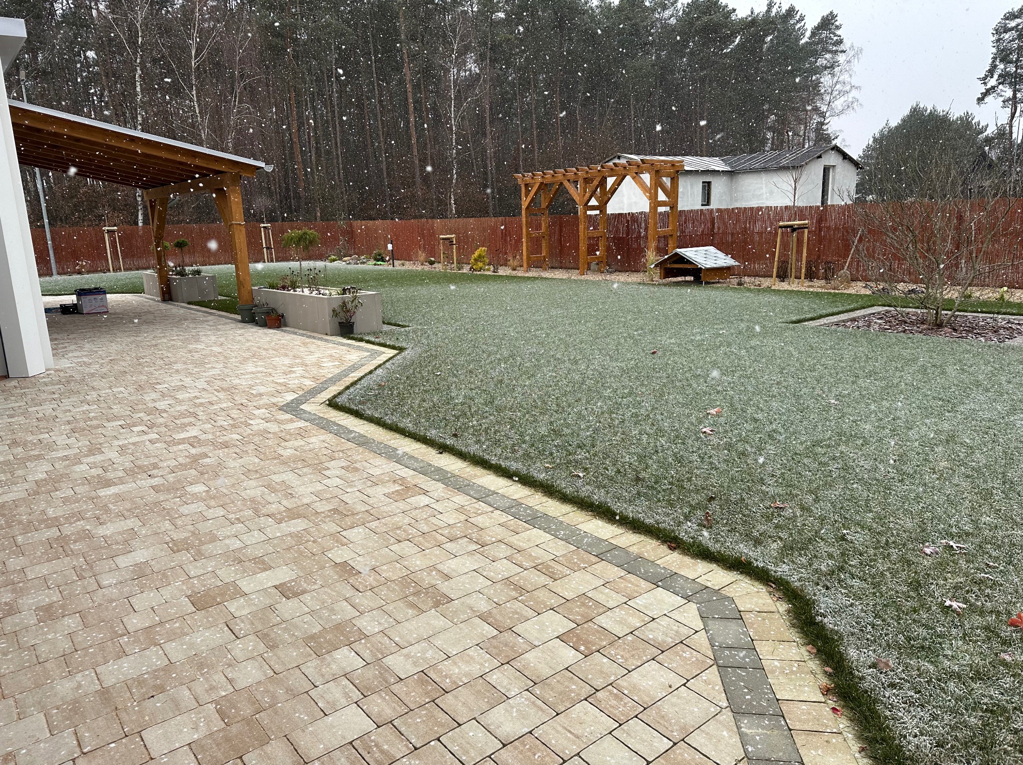 Zimowy ogród z kostką brukową, pergolą i świeżo założonym trawnikiem, delikatnie przyprószony śniegiem. Widoczne drzewa w tle i elementy małej architektury.