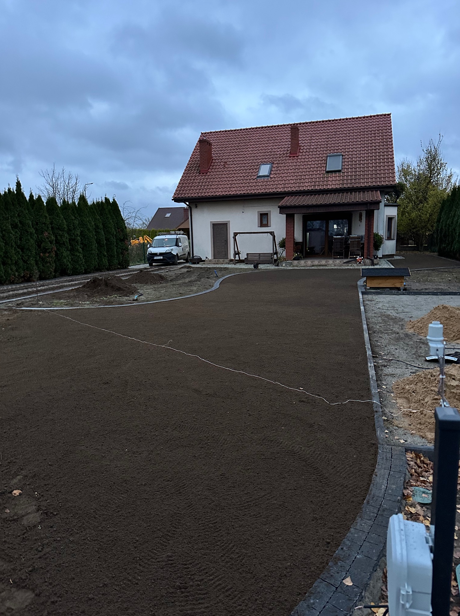Przygotowany teren pod trawnik z widocznym systemem nawadniania, obrzeżami i domem w tle. Świeżo nawieziona ziemia czeka na zasianie trawy.