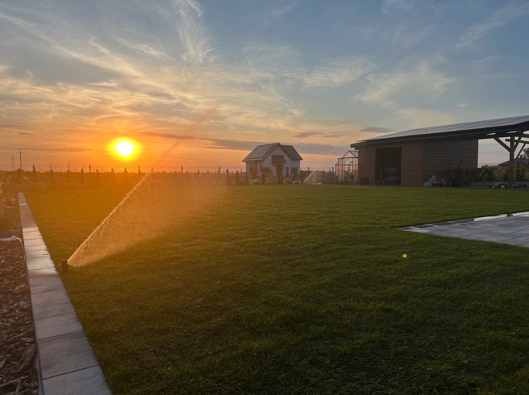 Automatyczne nawadnianie trawnika w Kłecku o zachodzie słońca. Zraszacze w akcji, w tle dom, szklarnia i wiata z panelami słonecznymi. Soczysta zieleń trawy.
