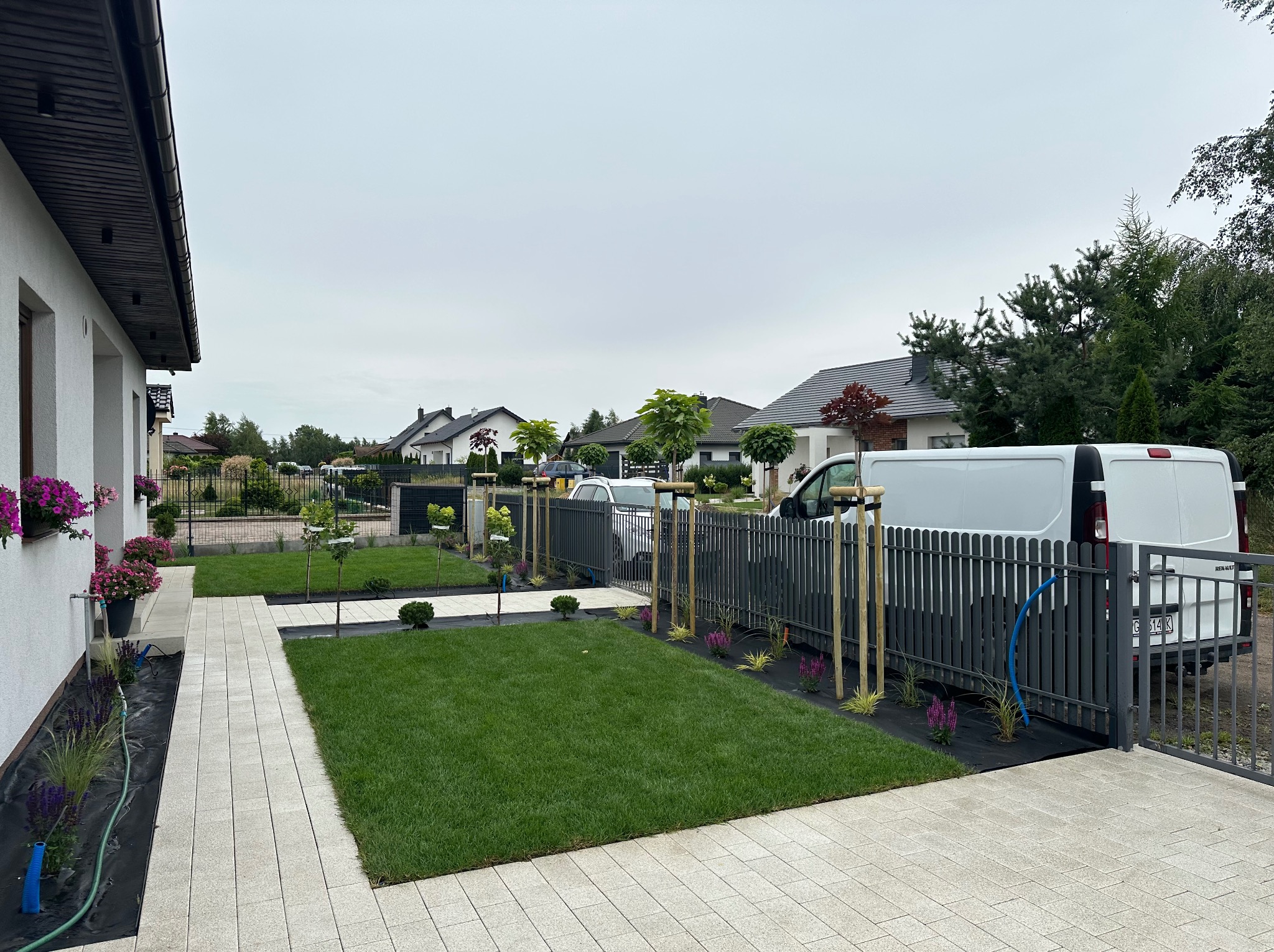 Nowoczesny ogród z równo przystrzyżonym trawnikiem, świeżo posadzone drzewka podparte palikami, rabaty kwiatowe i szara palisada. Na podjeździe stoi biały samochód dostawczy.