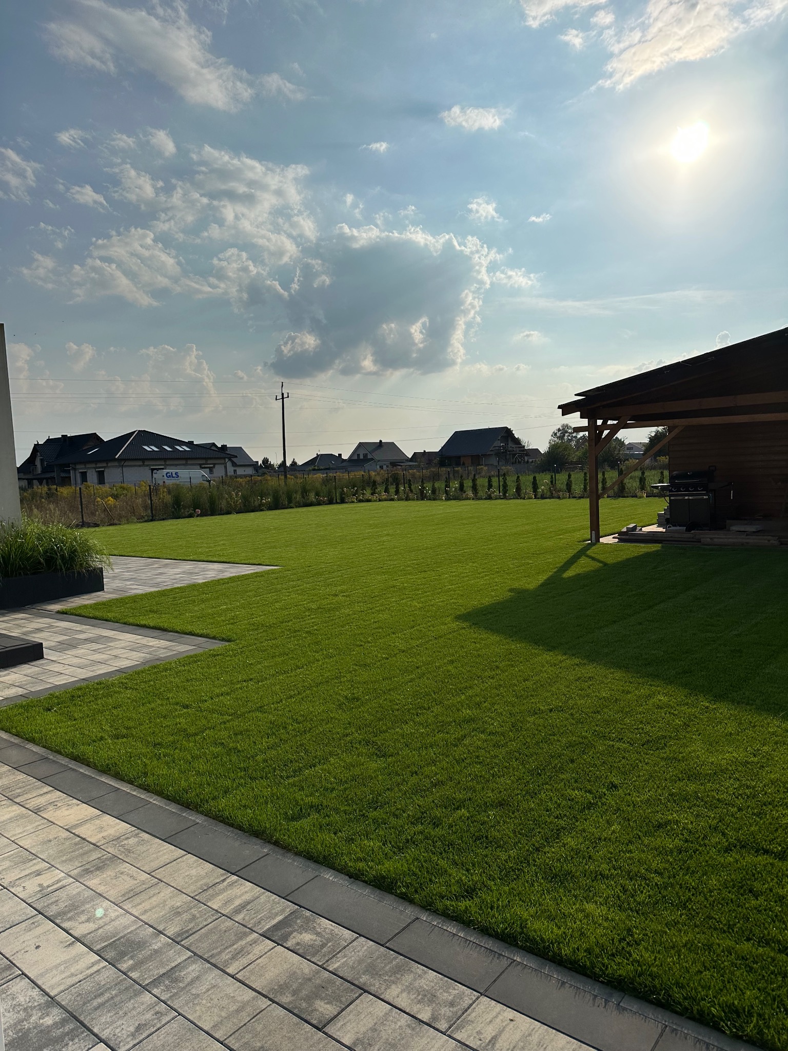 Perfekcyjnie utrzymany trawnik z rolki w słoneczny dzień, widok z poziomu kostki brukowej, zadaszenie grilla w tle, nowoczesne domy w oddali.