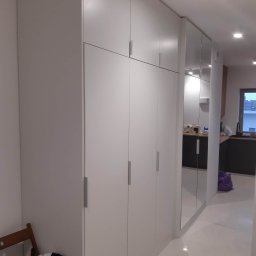 Michał Kamiński - Biała szafa wnękowa z lustrem, minimalistyczny design. Widoczne srebrne uchwyty i odbicie kuchni w lustrzanych drzwiach. Krzesło i torba obok szafy.