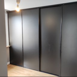Michał Kamiński - Elegancka szafa wnękowa z przesuwnymi, matowymi drzwiami w kolorze grafitowym, z delikatnym oświetleniem górnym i jasną podłogą z paneli.