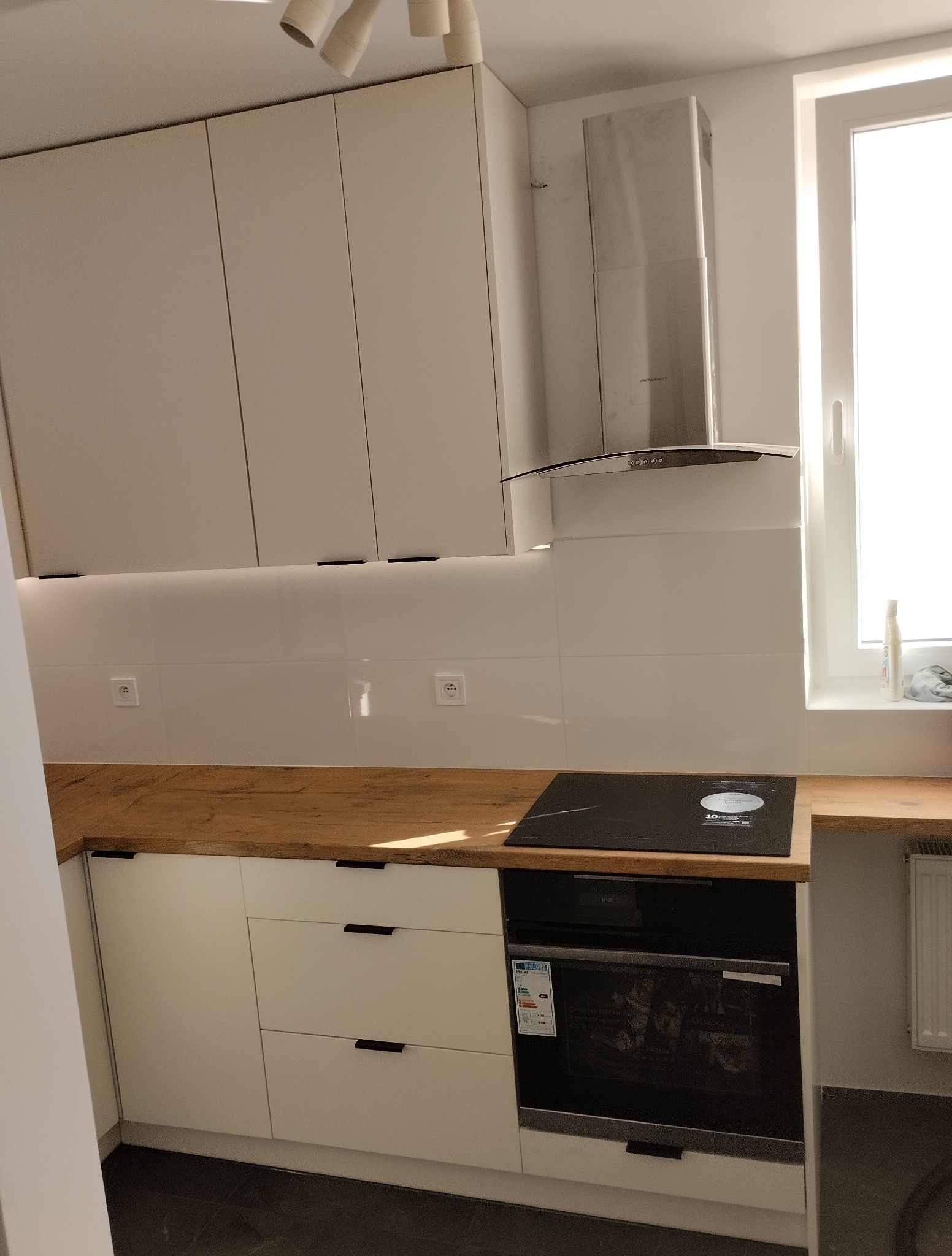 Jasna kuchnia z drewnianym blatem i białymi szafkami z minimalistycznymi, czarnymi uchwytami. Widoczna płyta indukcyjna i okap.