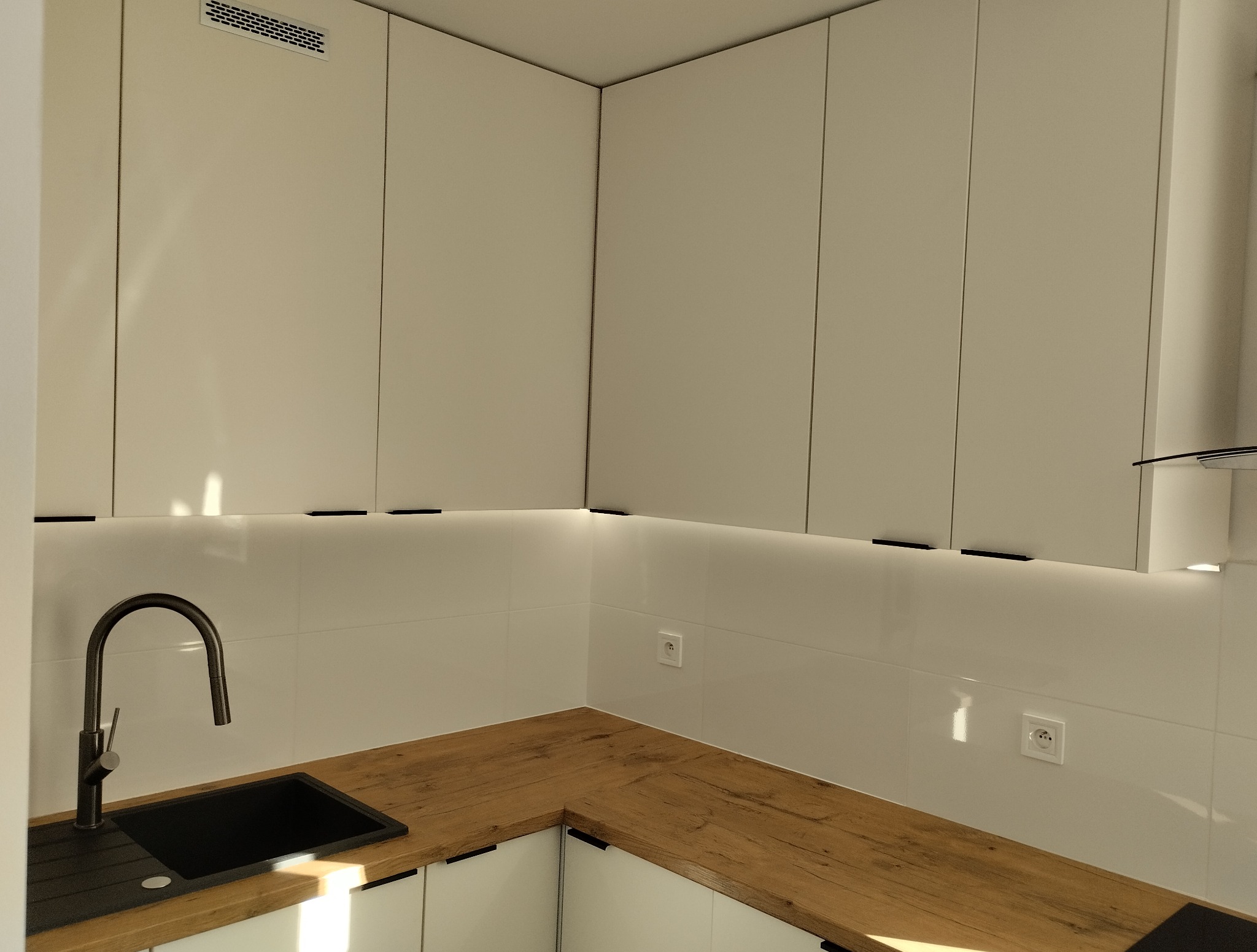 Nowoczesna kuchnia z białymi szafkami, drewnianym blatem i czarnym zlewem. Minimalistyczne uchwyty, białe płytki i delikatne oświetlenie LED pod szafkami.