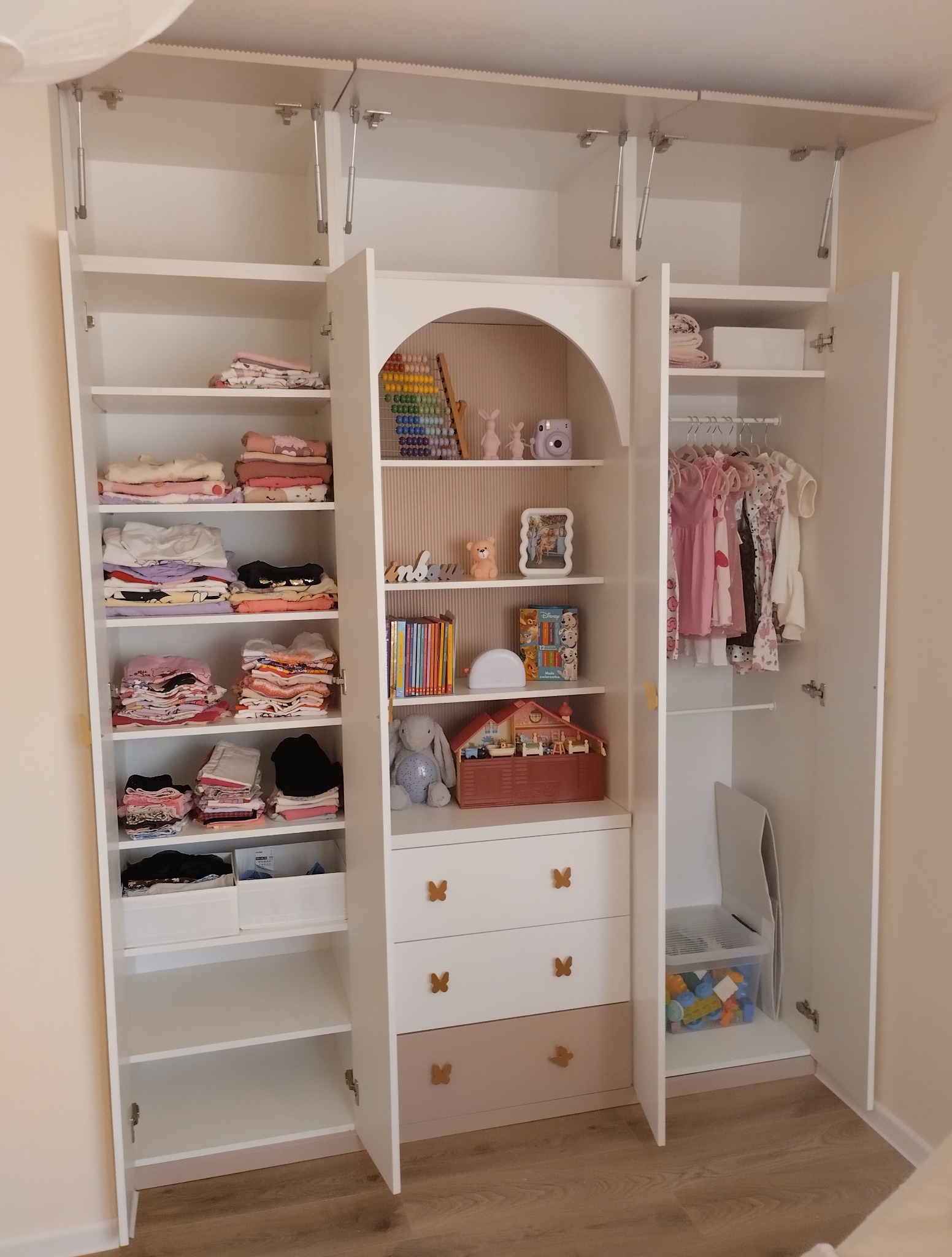 Biała szafa na wymiar dla dziecka z otwartymi drzwiami, półkami z ubraniami i zabawkami, z dekoracyjnymi uchwytami w kształcie motyli na szufladach.