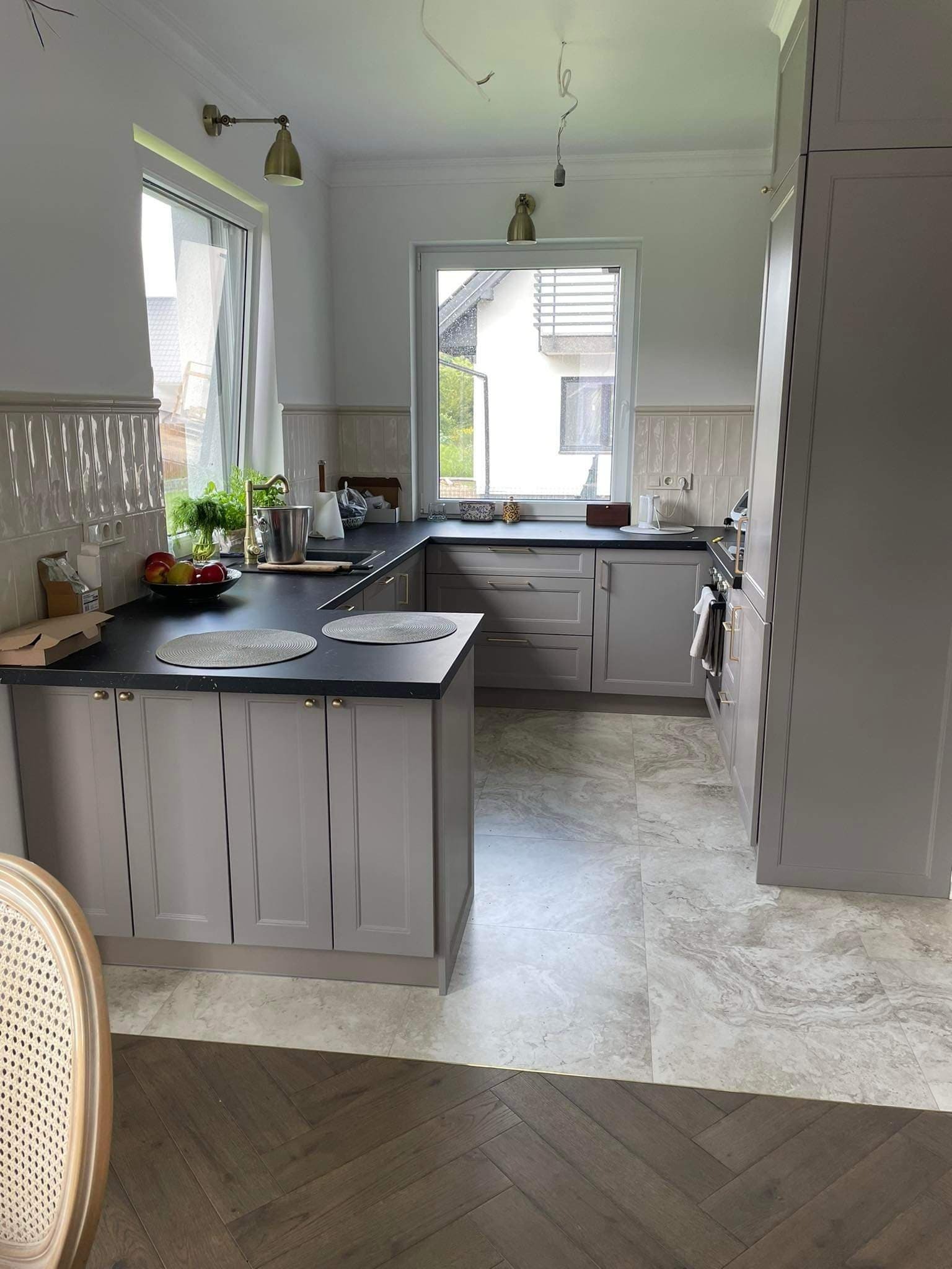 Jasnoszara kuchnia z czarnym blatem, widok na okno i dekoracyjne płytki ścienne. Uwagę przykuwają złote detale: bateria, uchwyty i lampy.