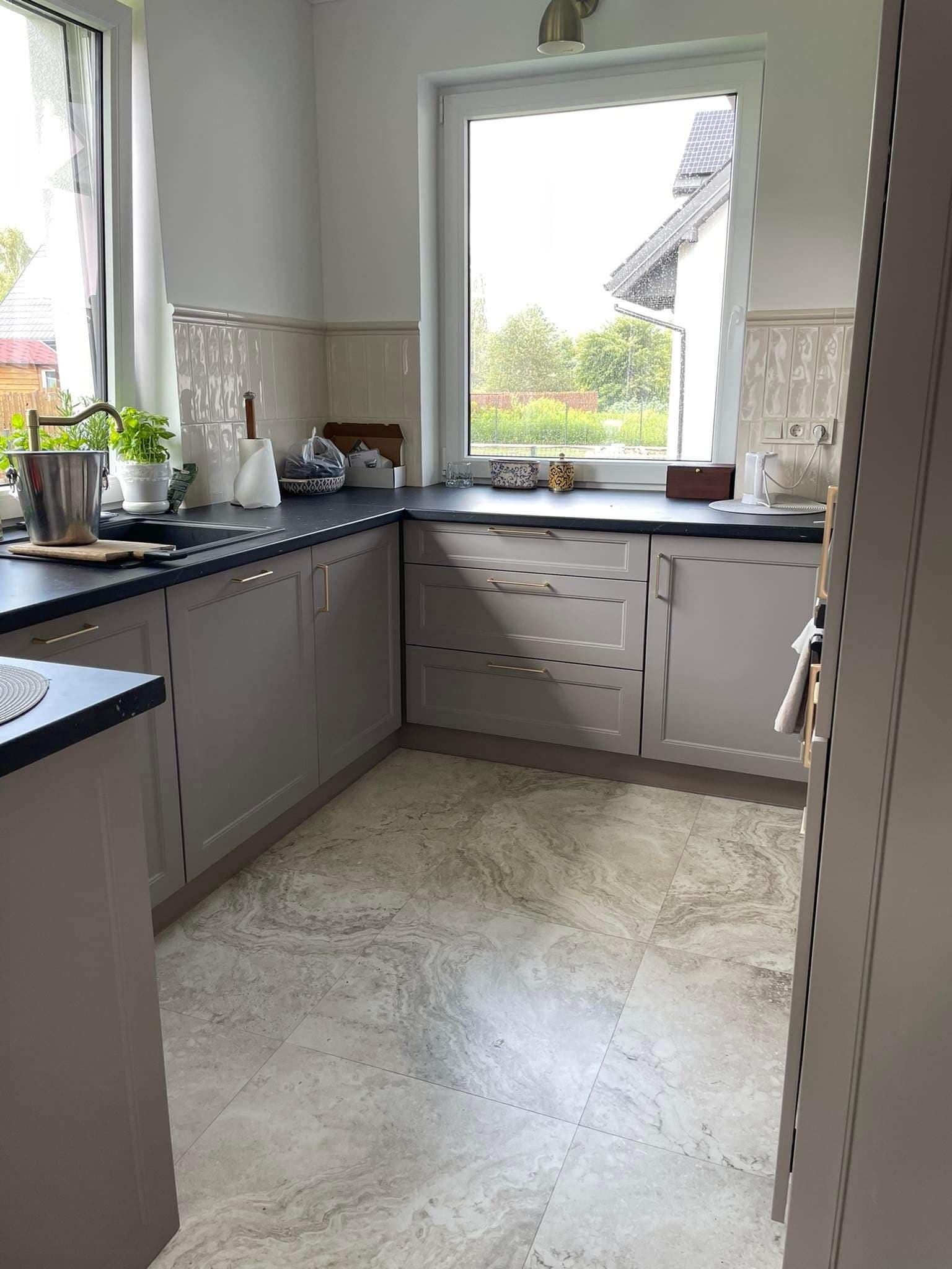 Jasna kuchnia z szarymi szafkami, czarnym blatem i marmurową podłogą. Widok na zewnątrz przez okno nad zlewem. Złote uchwyty dodają elegancji.
