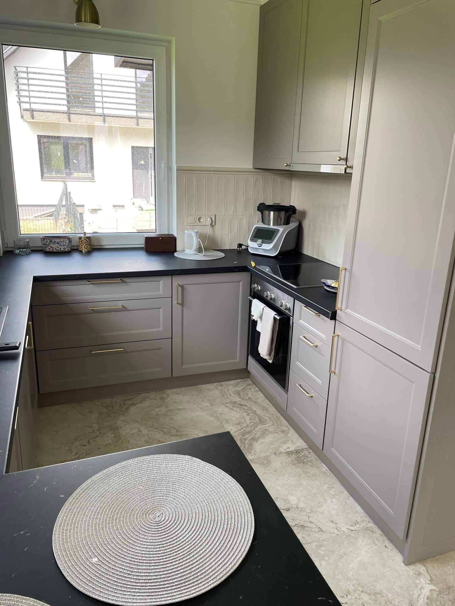Jasnoszara kuchnia z czarnym blatem, widok na okno i budynek z zewnątrz. Na blacie akcesoria kuchenne i dekoracyjny podkład. Marmurowe płytki na podłodze.