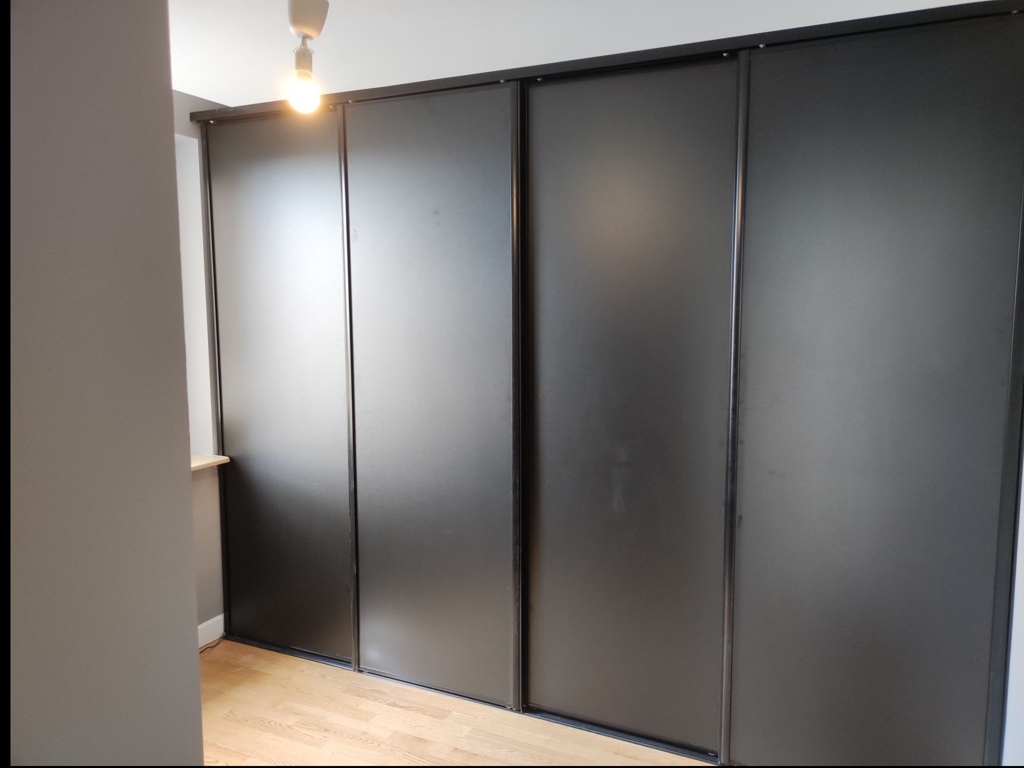 Elegancka szafa wnękowa z przesuwnymi, matowymi drzwiami w kolorze grafitowym, z delikatnym oświetleniem górnym i jasną podłogą z paneli.