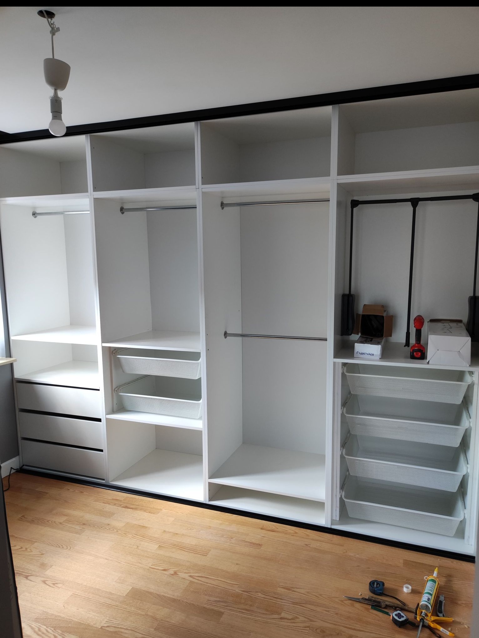Biała szafa wnękowa z czarnymi akcentami, otwarte półki, drążki na ubrania i wysuwane kosze. Na podłodze narzędzia montażowe.
