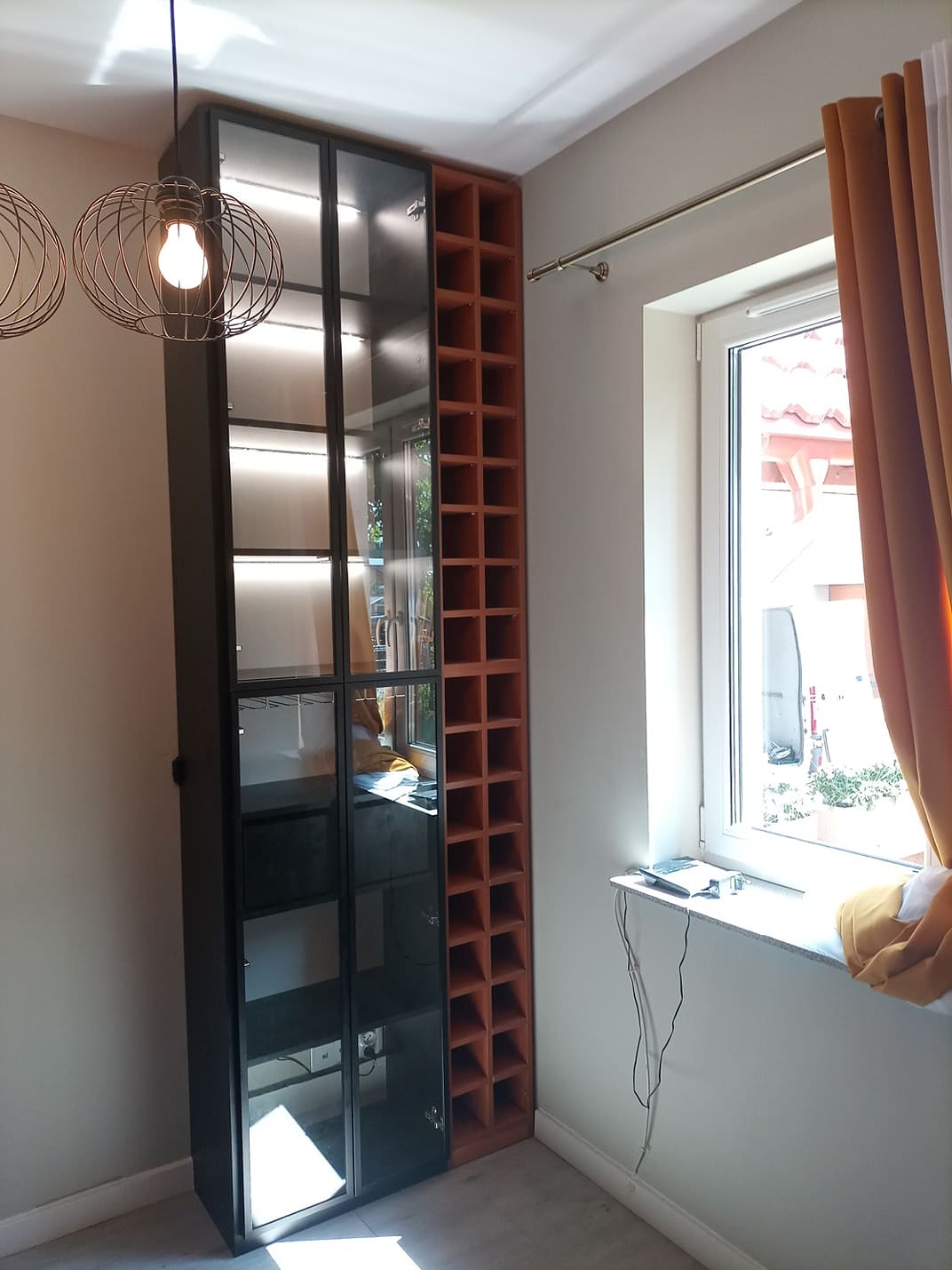 Nowoczesna szafa wnękowa z czarnymi ramami i szklanymi frontami, oświetlona od wewnątrz, sąsiadująca z regałem na wino w odcieniach brązu, w jasnym wnętrzu.