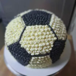 Tort w kształcie piłki nożnej, kremowy, dwukolorowy (biało-czarny), dekorowany w stylu cukierniczym na białym talerzu. Precyzyjne wykonanie.