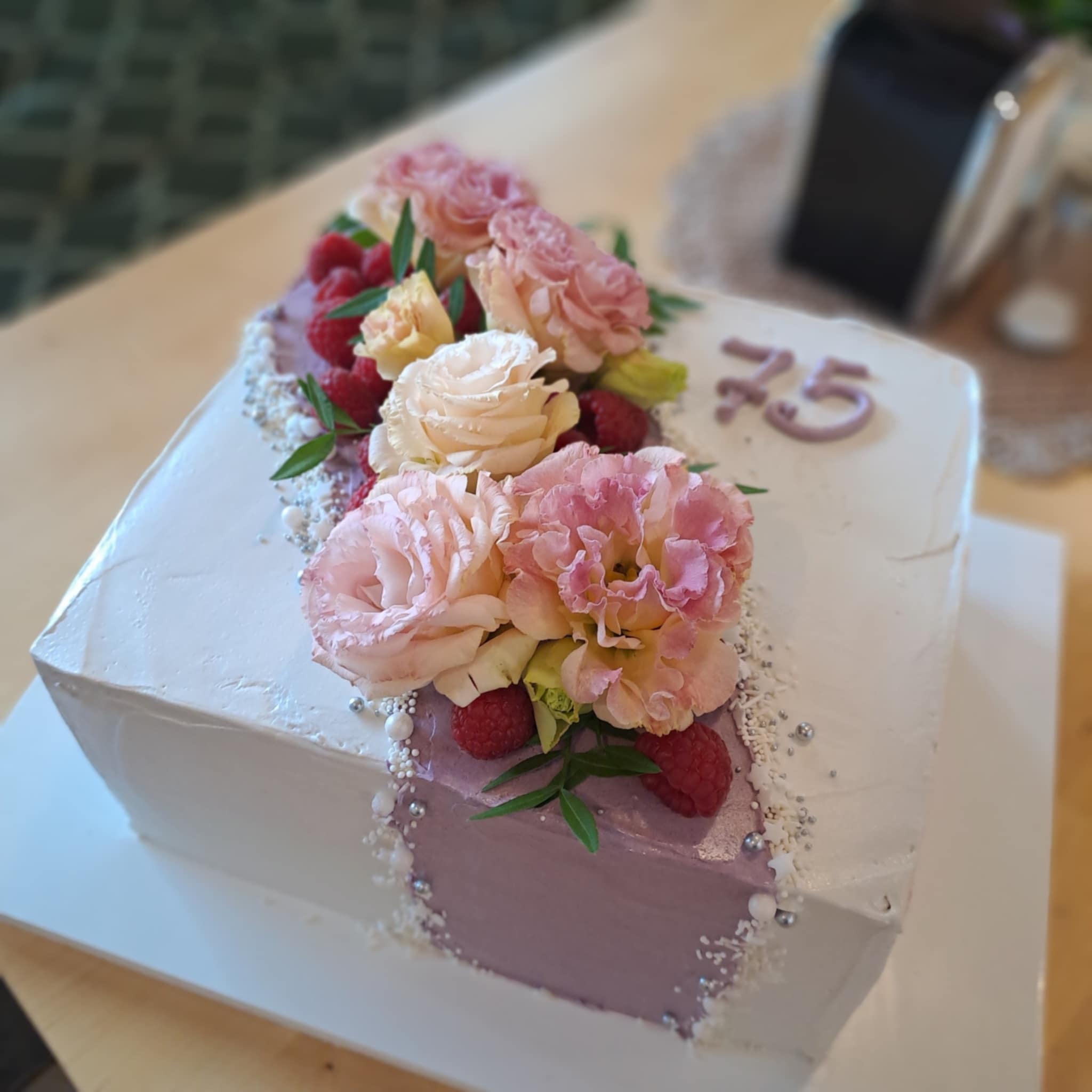 Kwadratowy tort na 75 urodziny, zdobiony świeżymi kwiatami w odcieniach różu i kremu, malinami i srebrnymi perełkami. Kremowy lukier i fioletowy akcent.