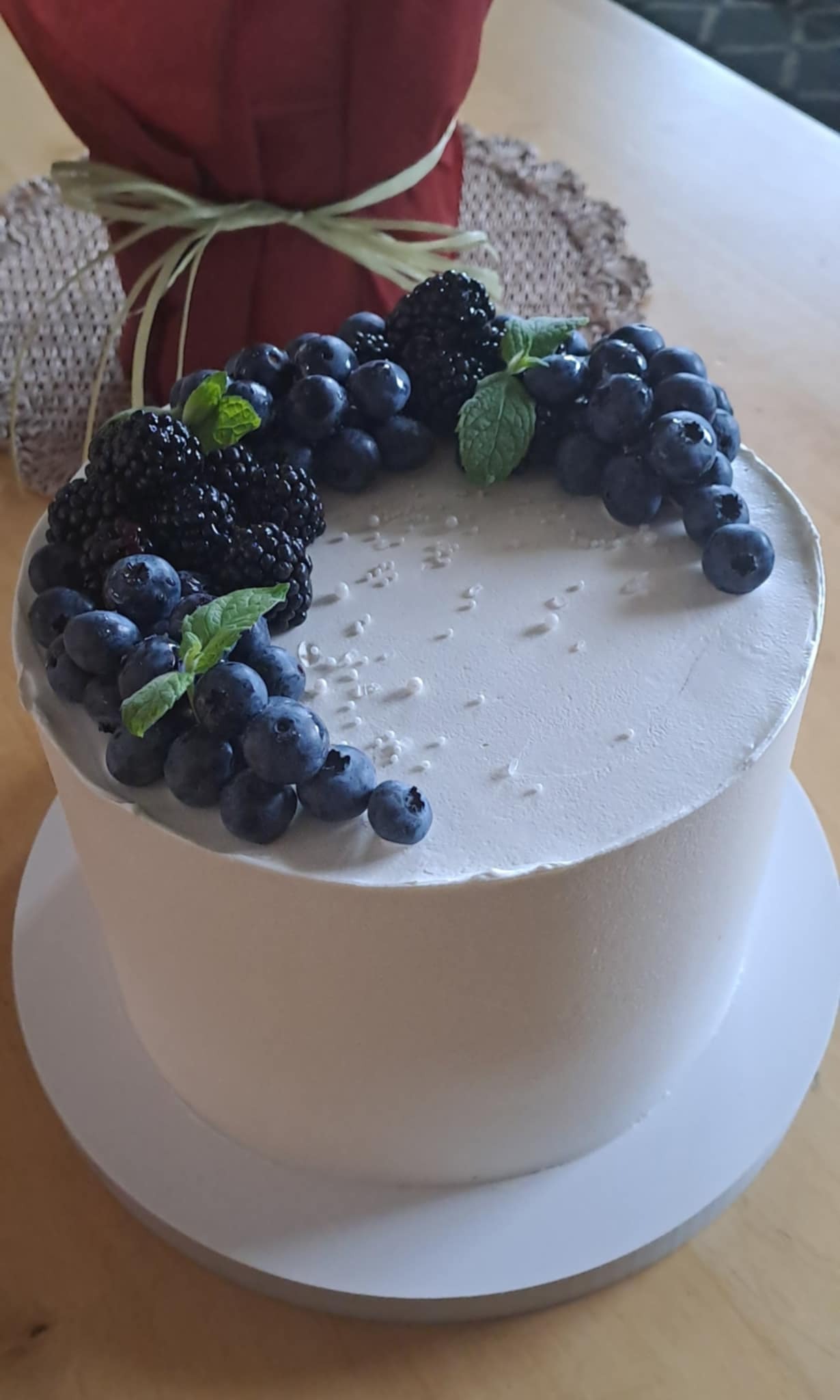 Tort z białym kremem, dekorowany świeżymi borówkami, jeżynami i listkami mięty. Tło stanowi dekoracja stołu z czerwoną tkaniną.