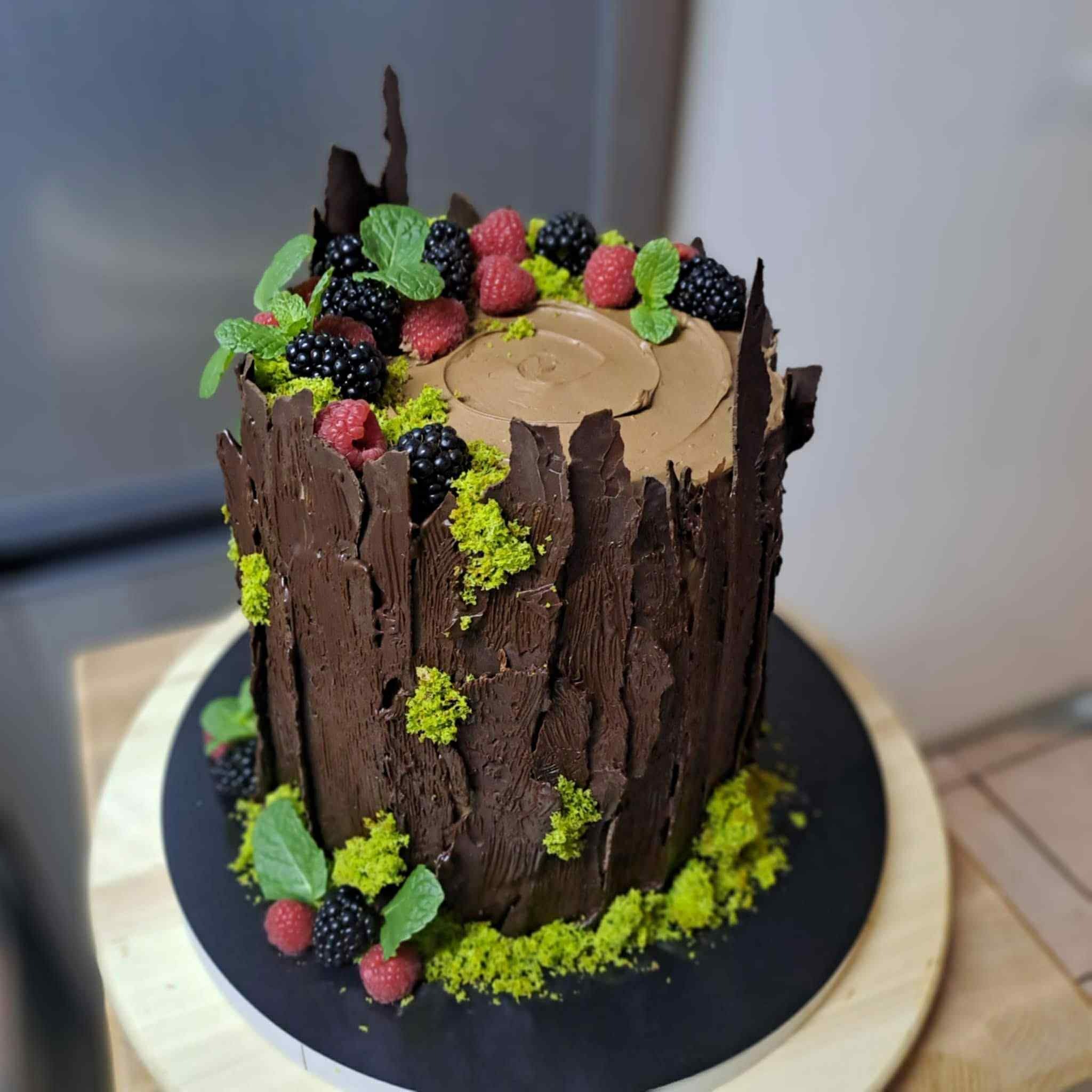 Tort w kształcie pnia drzewa, pokryty czekoladą imitującą korę, udekorowany malinami, jeżynami, miętą i zielonymi okruchami, na czarnym talerzu.