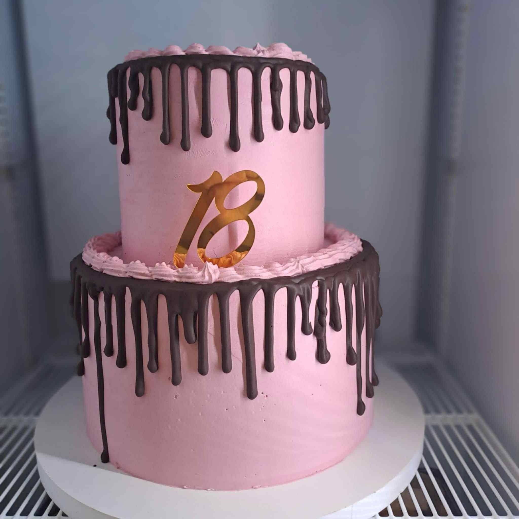 Dwupiętrowy tort w odcieniach różu z czekoladowymi polewami i złotą dekoracją '18', umieszczony na białym stojaku w chłodnym otoczeniu.