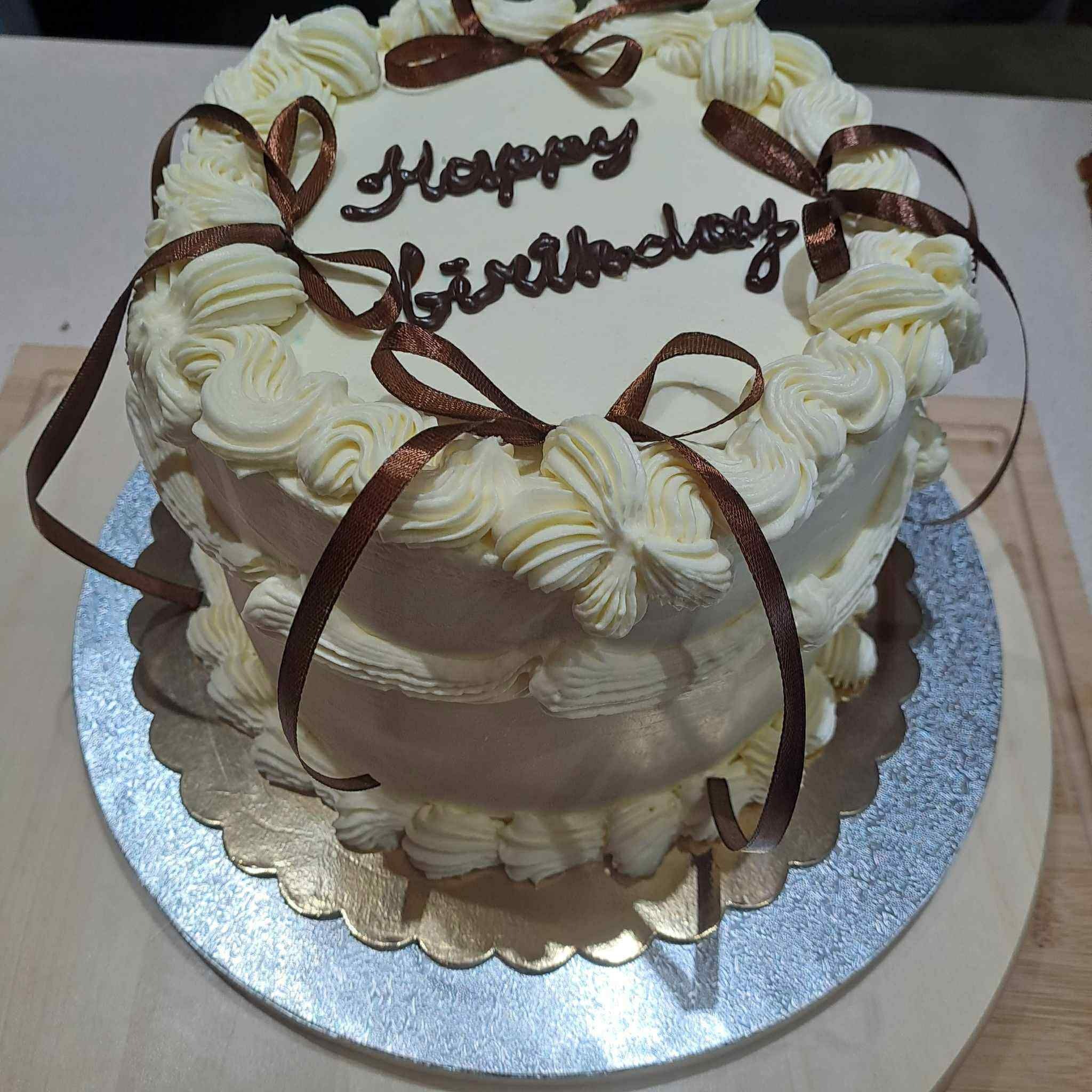 Tort urodzinowy z kremem, czekoladowym napisem i brązowymi wstążkami, umieszczony na srebrnej podkładce na drewnianym blacie. Dekoracja cukiernicza.
