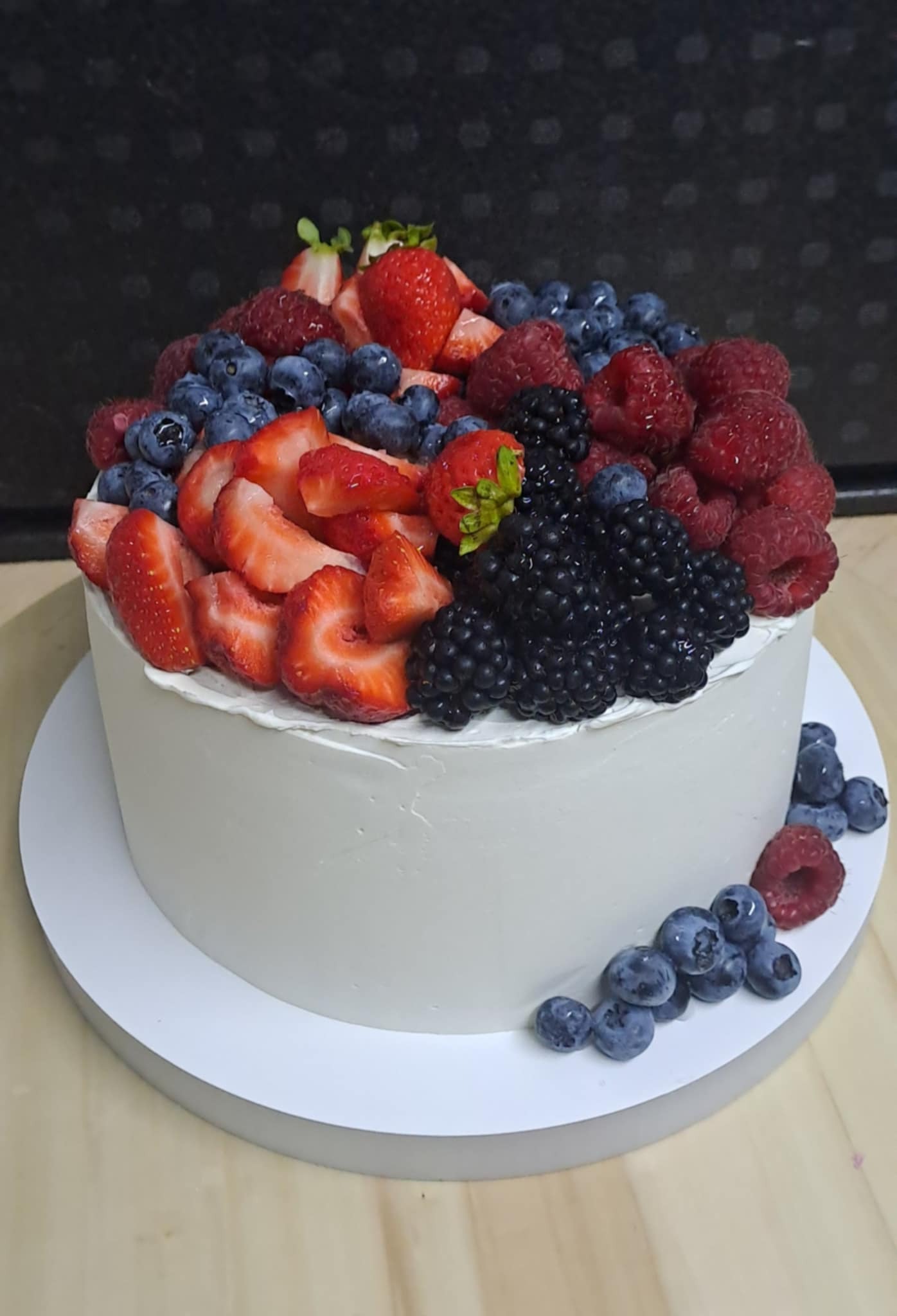 Tort z białym kremem, udekorowany świeżymi owocami: truskawkami, malinami, jeżynami i borówkami, ułożonymi w rzędach. Widoczne pojedyncze owoce na podstawie tortu.