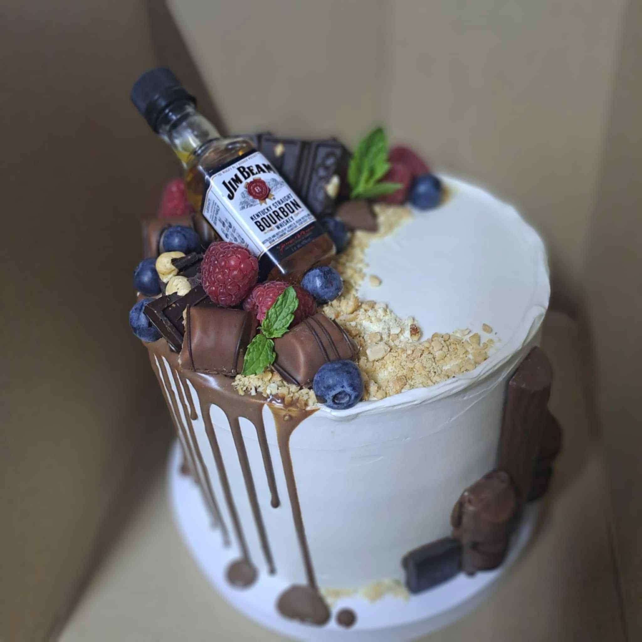 Tort z białym kremem, polewą czekoladową, owocami (maliny, borówki), kawałkami czekolady i buteleczką Jim Beam. Dekoracja na urodziny.