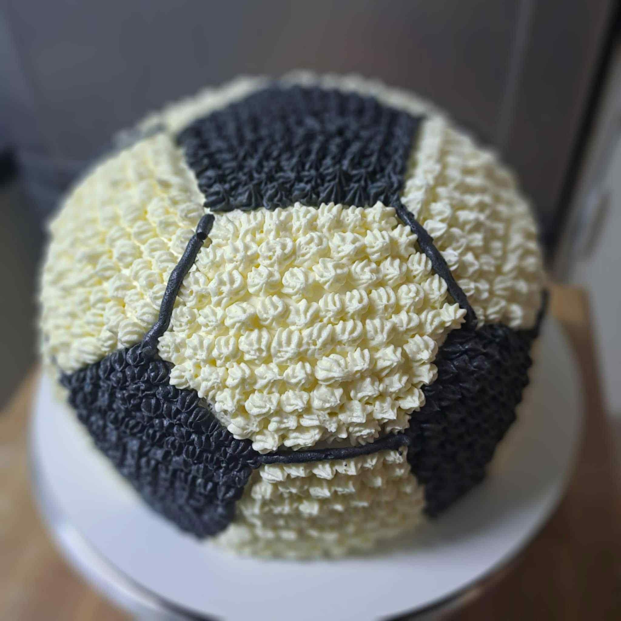 Tort w kształcie piłki nożnej, kremowy, dwukolorowy (biało-czarny), dekorowany w stylu cukierniczym na białym talerzu. Precyzyjne wykonanie.