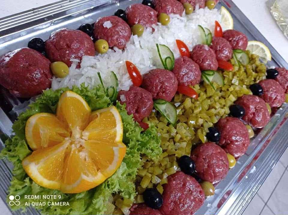 Elegancko podany tatar wołowy na srebrnej tacy, z dekoracją z pomarańczy, sałaty, ogórków, oliwek i białej rzodkwi, tworzący apetyczną kompozycję.