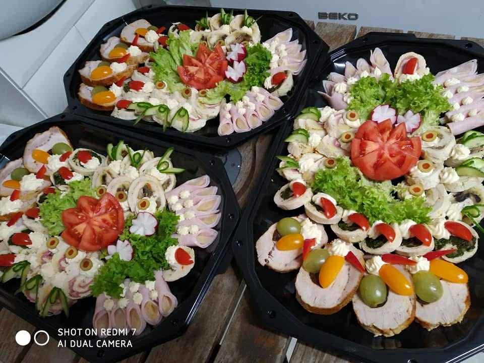 Trzy czarne tace z zimnymi przekąskami: roladki z szynki, koreczki, kanapki dekorowane warzywami. Centralnie ułożone pomidory i sałata. Widoczny blat drewnianego stołu.